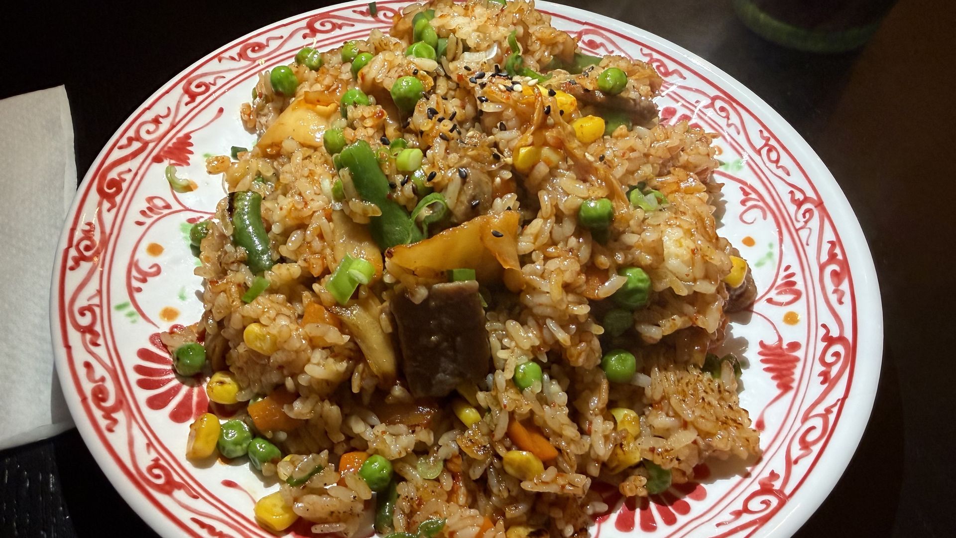 White plate with a red decorative rim, piled high with fried rice containing peas, corn, carrots, and beef chunks, topped with sesame seeds and chopped green onions.