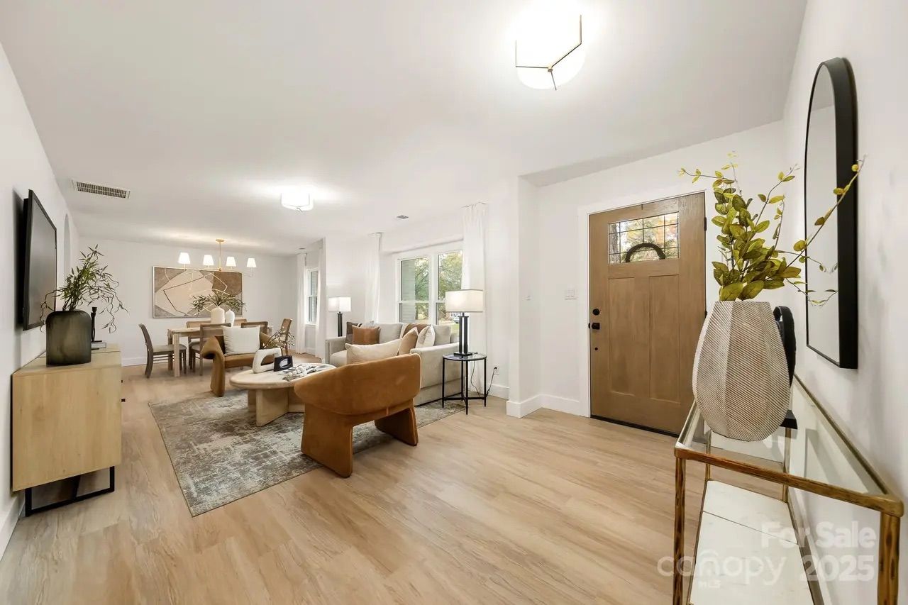 Bright open living room with light wood floor, beige and brown furniture, modern dining table, abstract wall art, and wooden front door with glass panel and a wreath.