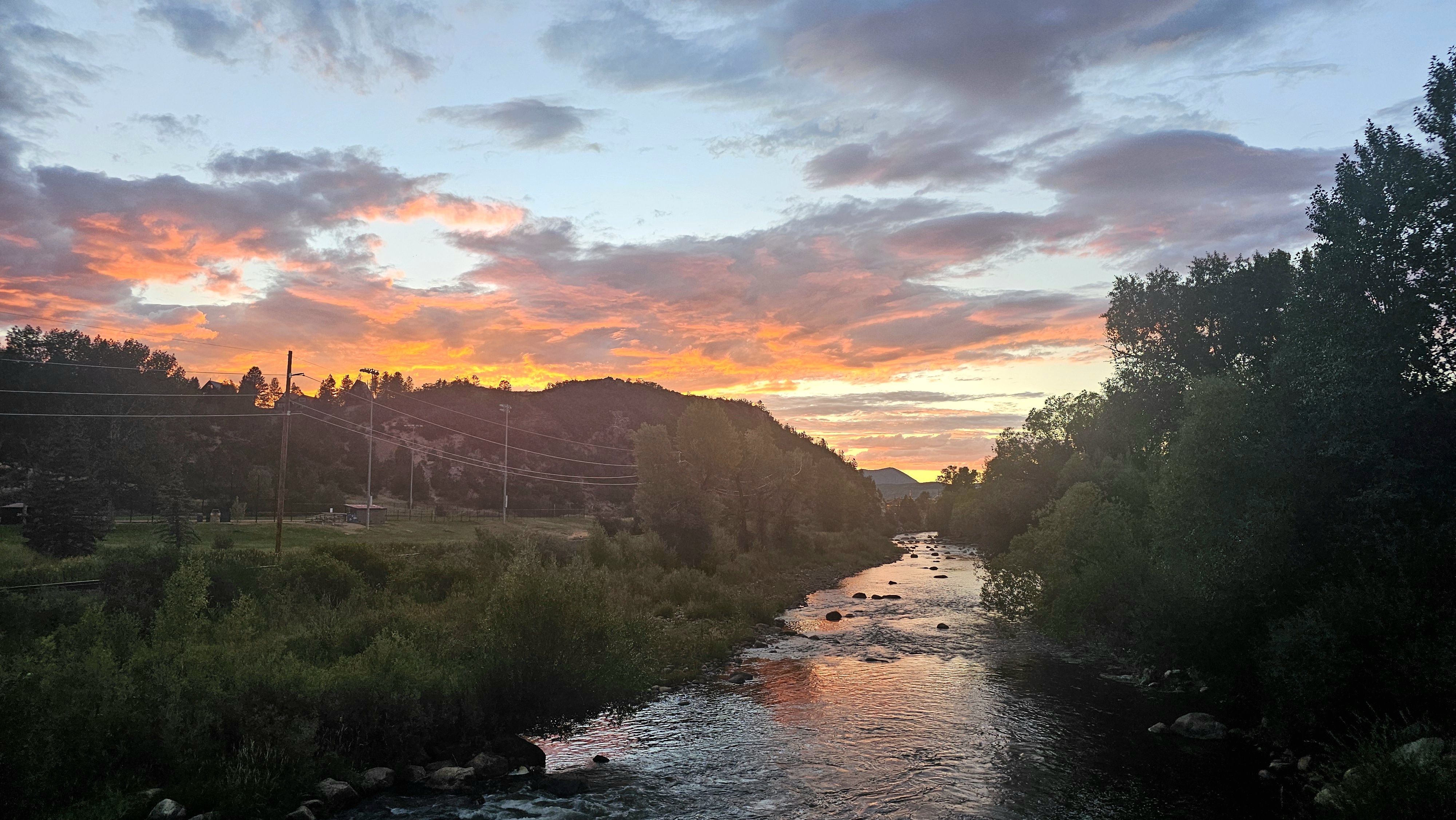 Sunset over a river flanked by tree-lined banks and distant hills, with orange-pink clouds lighting the sky; power poles and lines run along a road on the left.