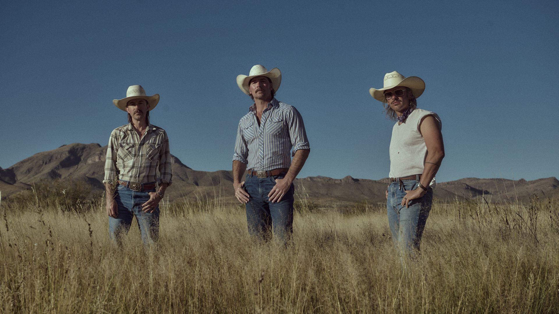 Three cowboys in wide-brim hats stand in a dry, tall field with arid mountains under a clear blue sky. They wear denim jeans and plaid or striped shirts, facing the camera.