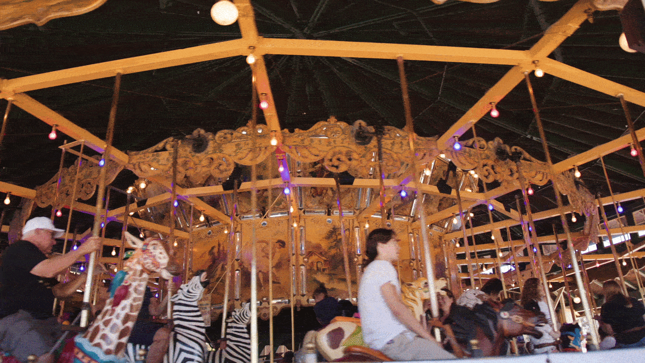 Indoor carousel with a wooden frame and ornate top, colorful string lights, and riders on carved horses. A man in a cap sits left while others in zebra-pattern outfits ride around.