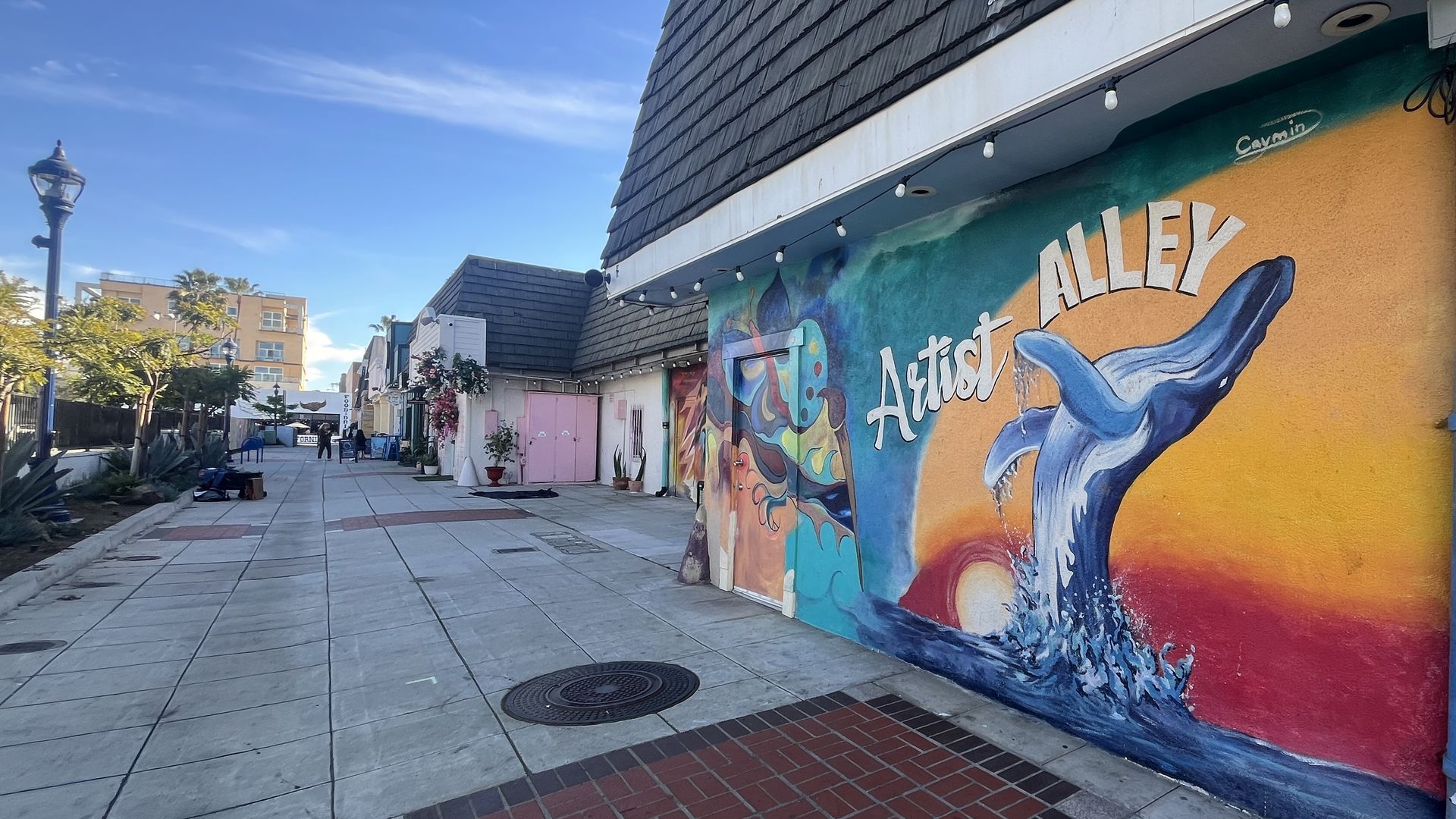 Sunny alley with a mural on the right wall reading "Artist Alley" in white over teal and orange sunset, featuring a blue whale splashing out of the ocean. 