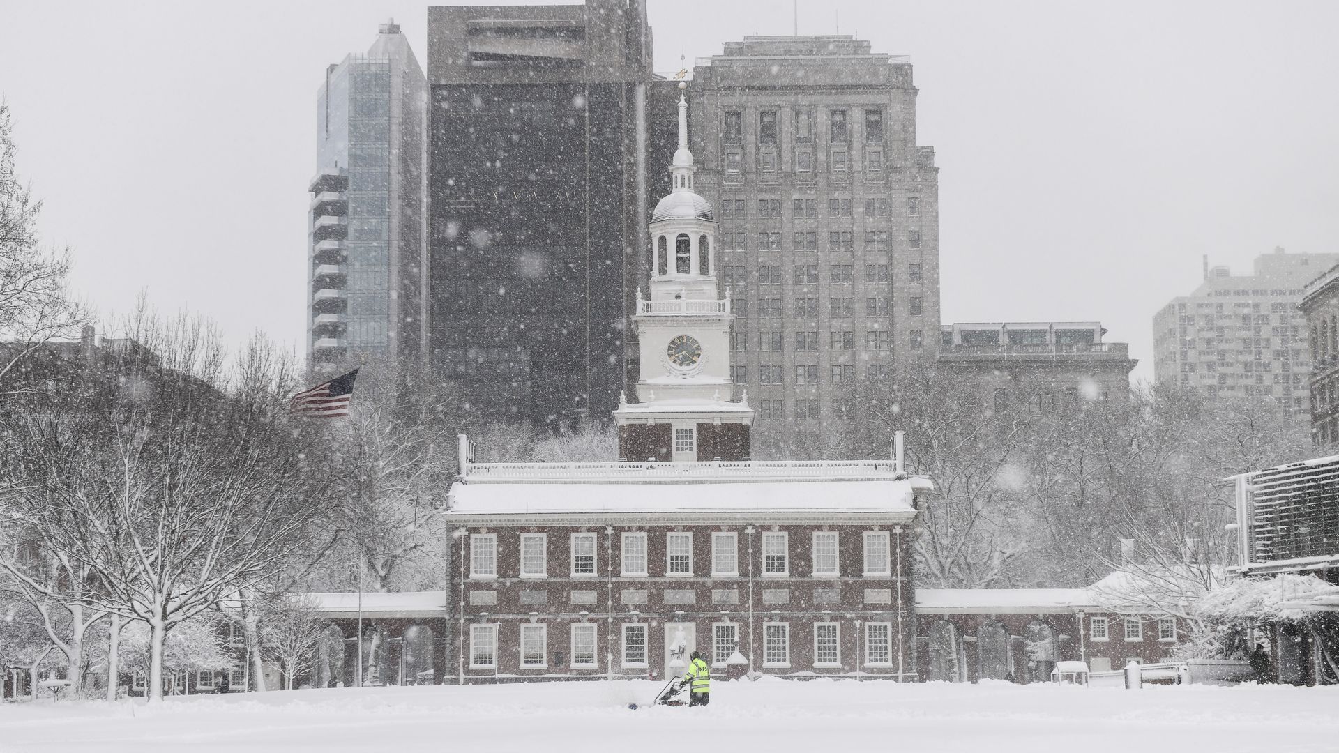 Snow-covered Independence Hall in Philadelphia with person in bright jacket clearing snow in heavy snowfall, bare trees, and tall buildings in background under gray sky.