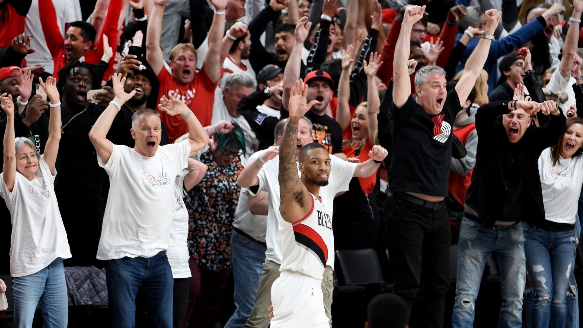 Basketball player in white Portland Trail Blazers uniform raises hand amid cheering crowd with raised arms and excited expressions during a game.