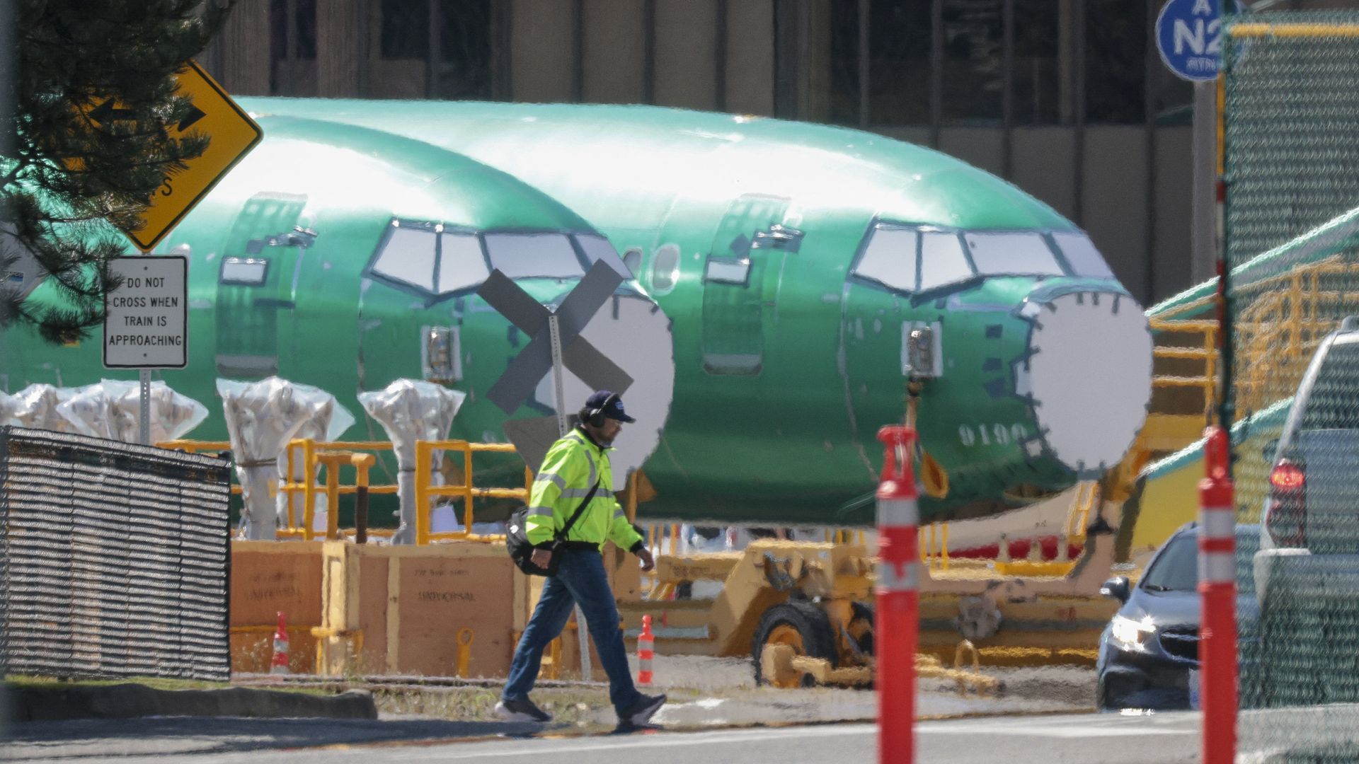 Person in a neon yellow jacket and headphones walking past two green airplane fuselages under construction, with safety cones and signs around a fenced industrial area.