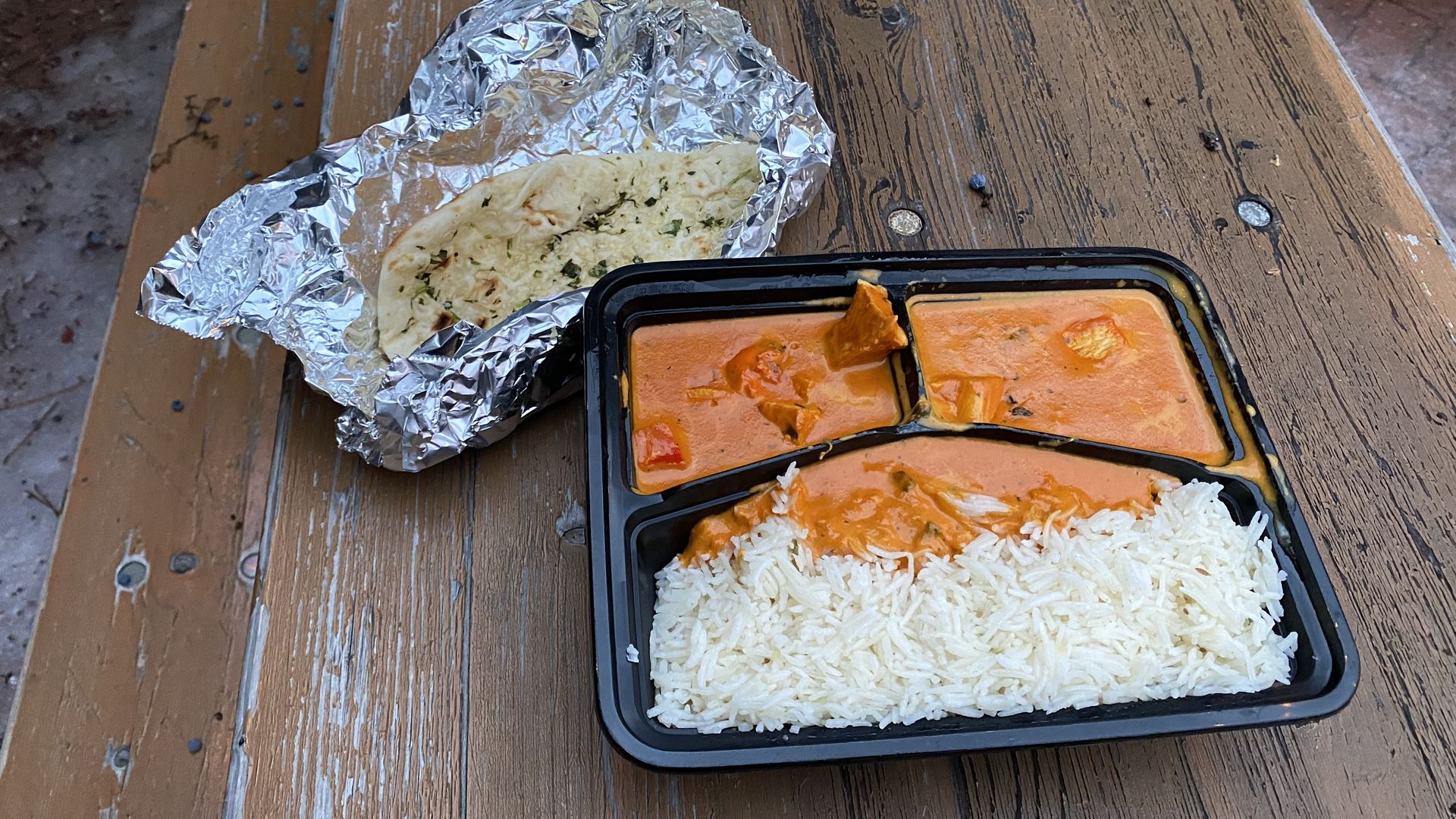 A black plastic container with three sections full of rice and chicken tikka masala, next to garlic naan in foil