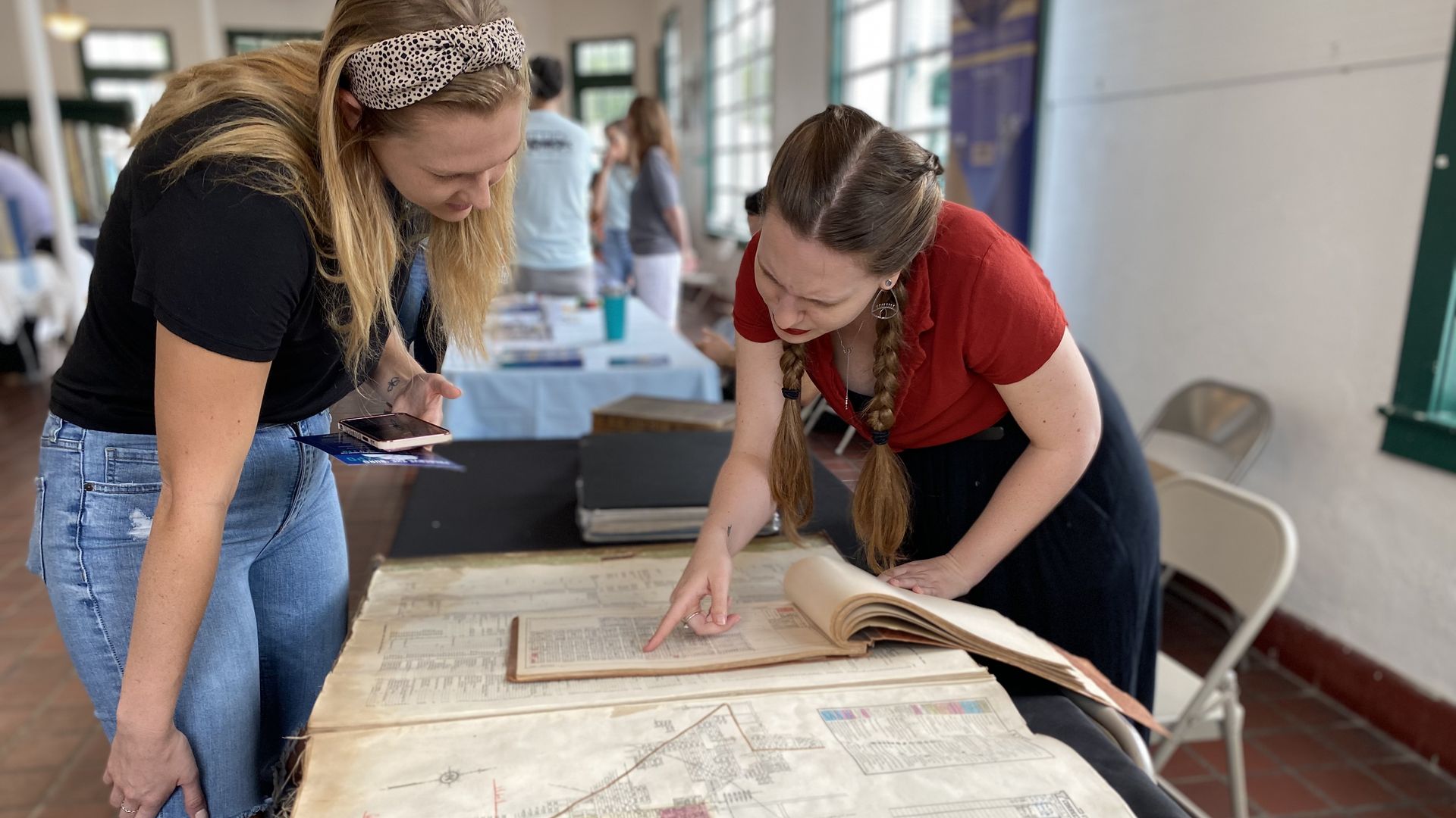 Two women lean over a large vintage atlas on a table, studying maps. One wears a black shirt and jeans with a spotted headband; the other wears a red shirt with braided hair in a bright room.