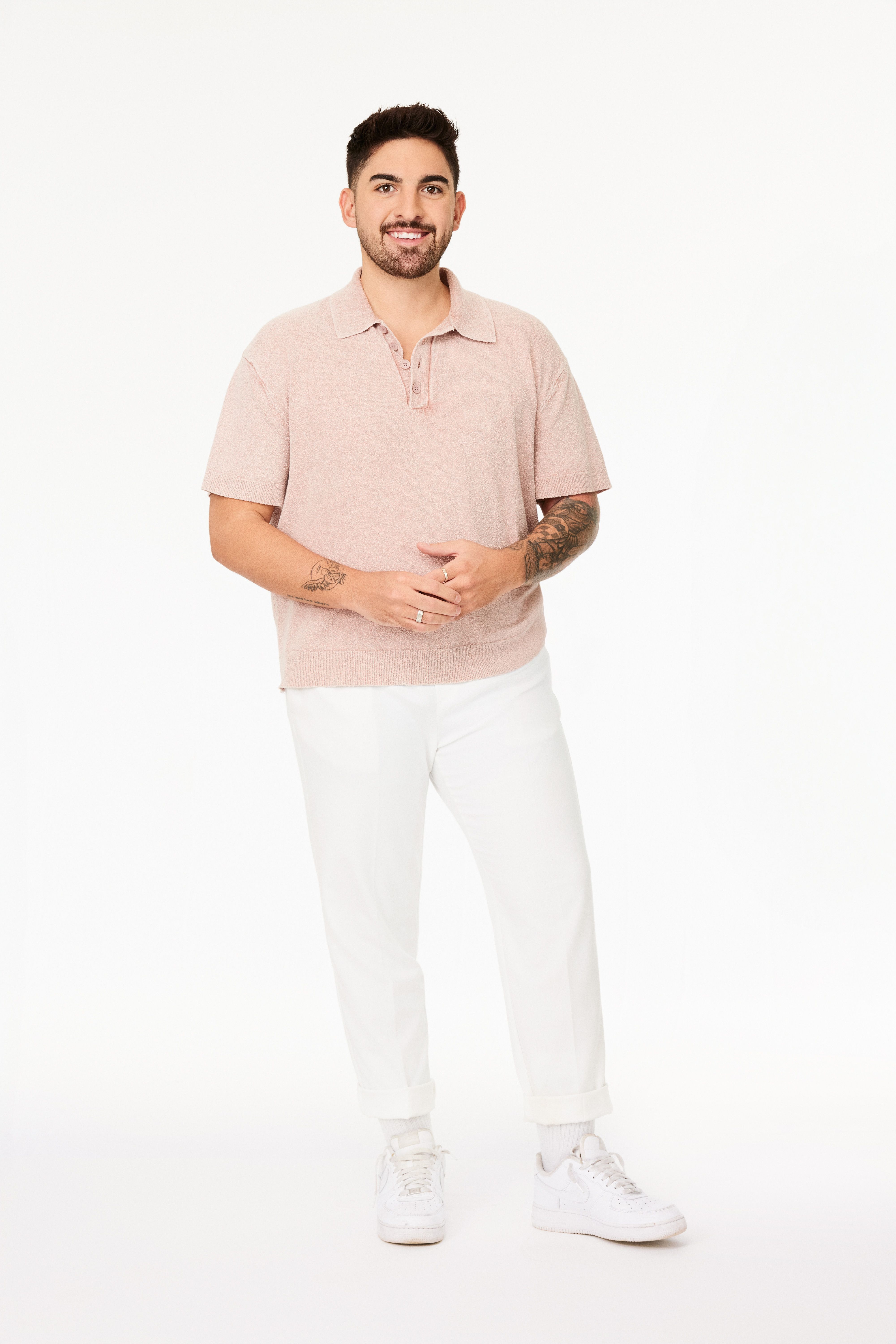 Man with short dark hair and beard smiling, wearing a light pink short-sleeve polo, white pants, white sneakers, and showing tattoos on both forearms, standing against a white background.