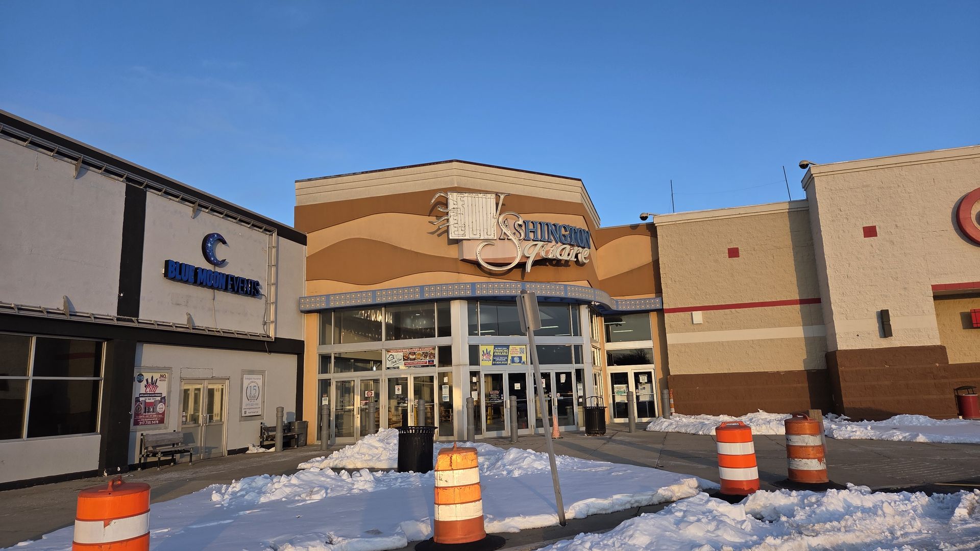 Exterior of Washington Square shopping center with blue sky, snow-covered ground, orange and white traffic barrels, and a sign for Blue Moon Events on the left.