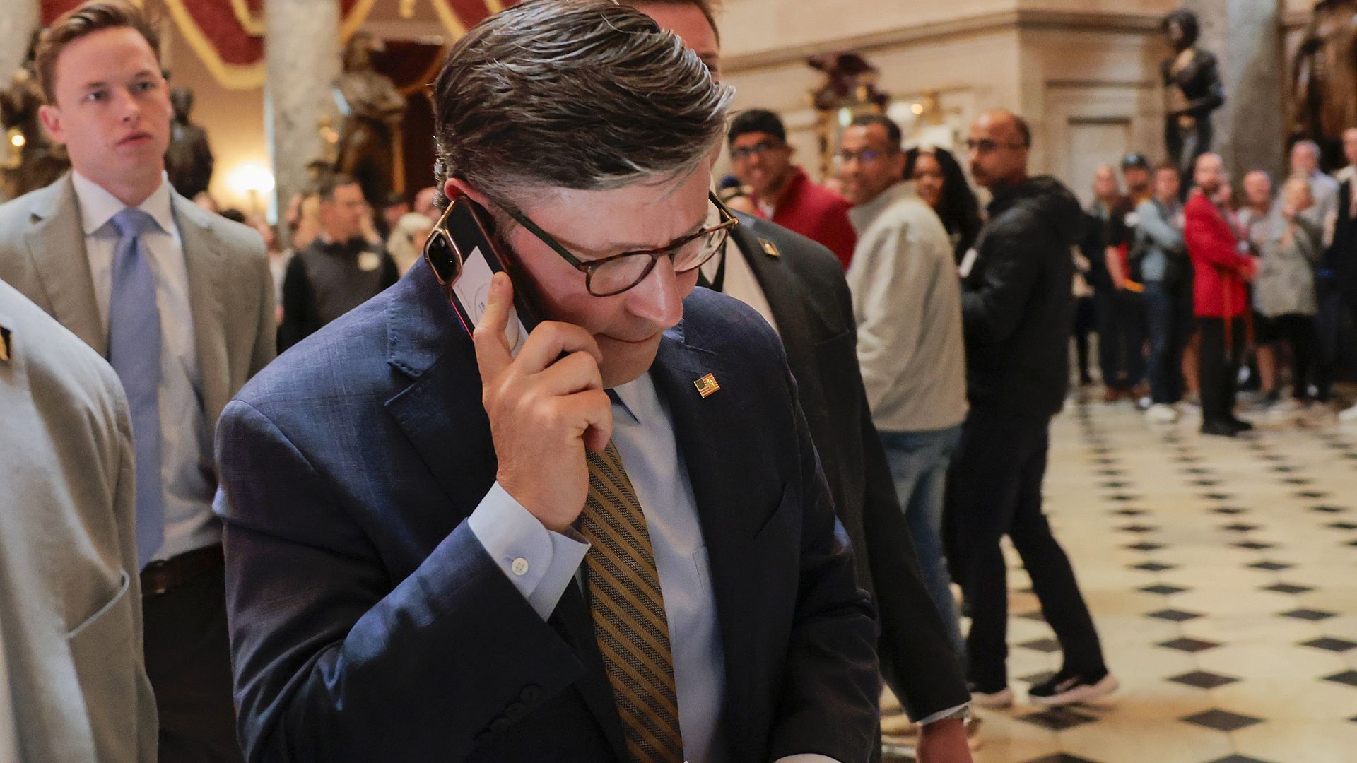 Man in a dark blue suit and glasses talks on a cellphone at a crowded indoor event, holding documents. A line of people and a checkered marble floor fill the background.