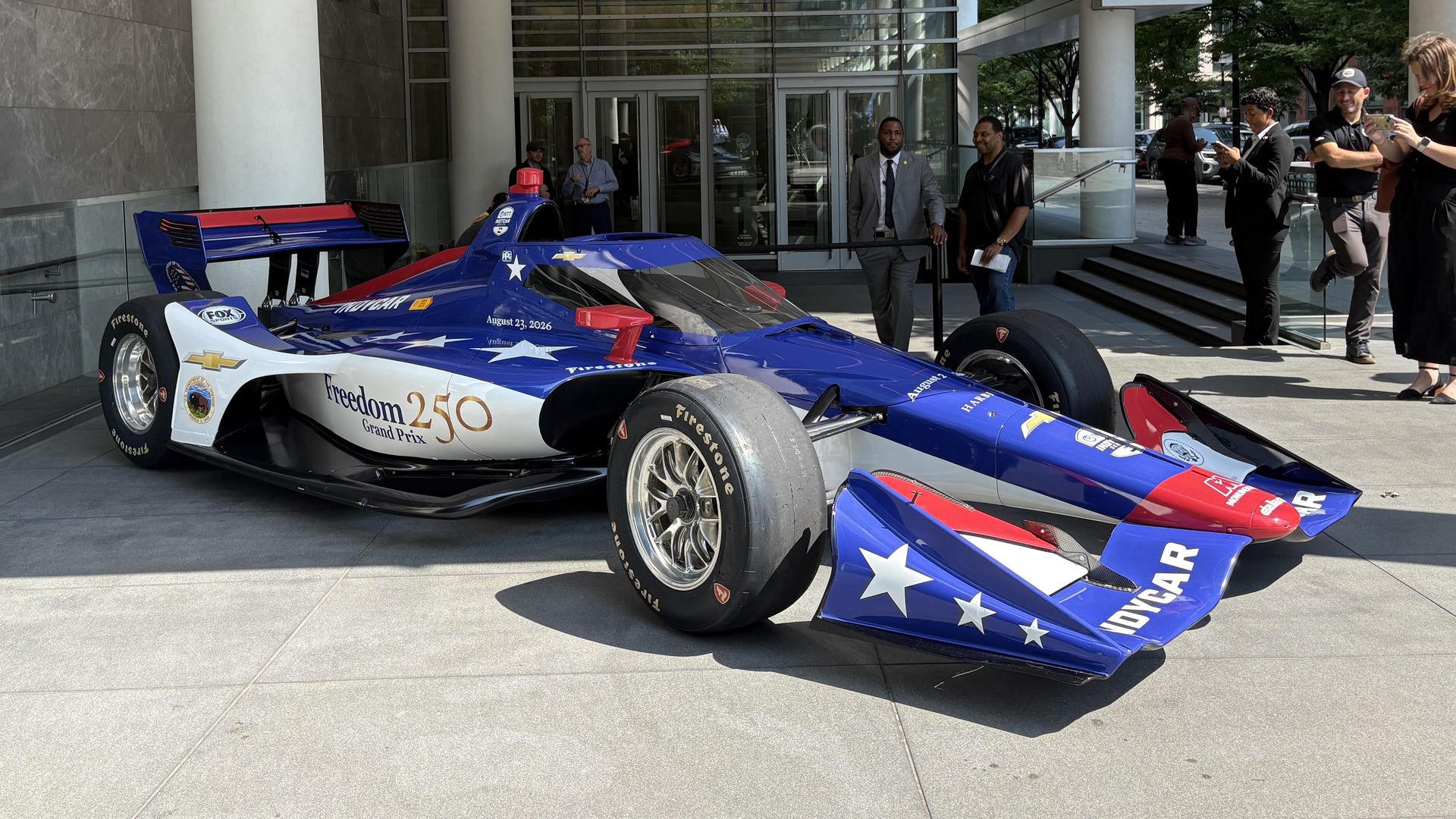 Outdoor scene at a modern glass building entrance with tall white columns. A blue IndyCar race car with white stars and the text "Freedom 250 Grand Prix" is parked, while several people stand near the doors taking photos.
