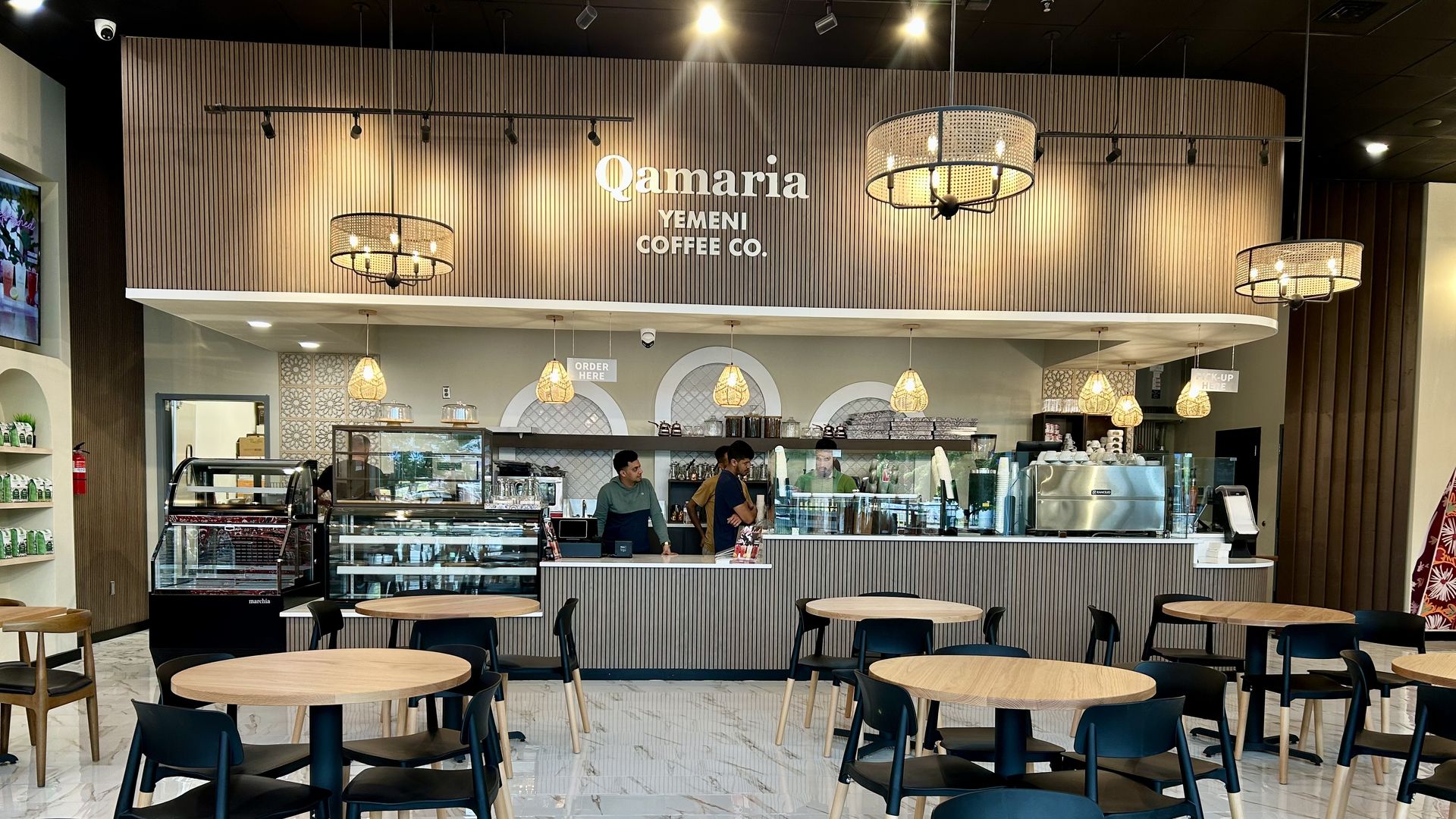 Interior of Qamaria Yemeni Coffee Co. with wood-paneled walls, hanging lights, round wooden tables, black chairs, and a marble-patterned floor. Baristas stand behind the counter.