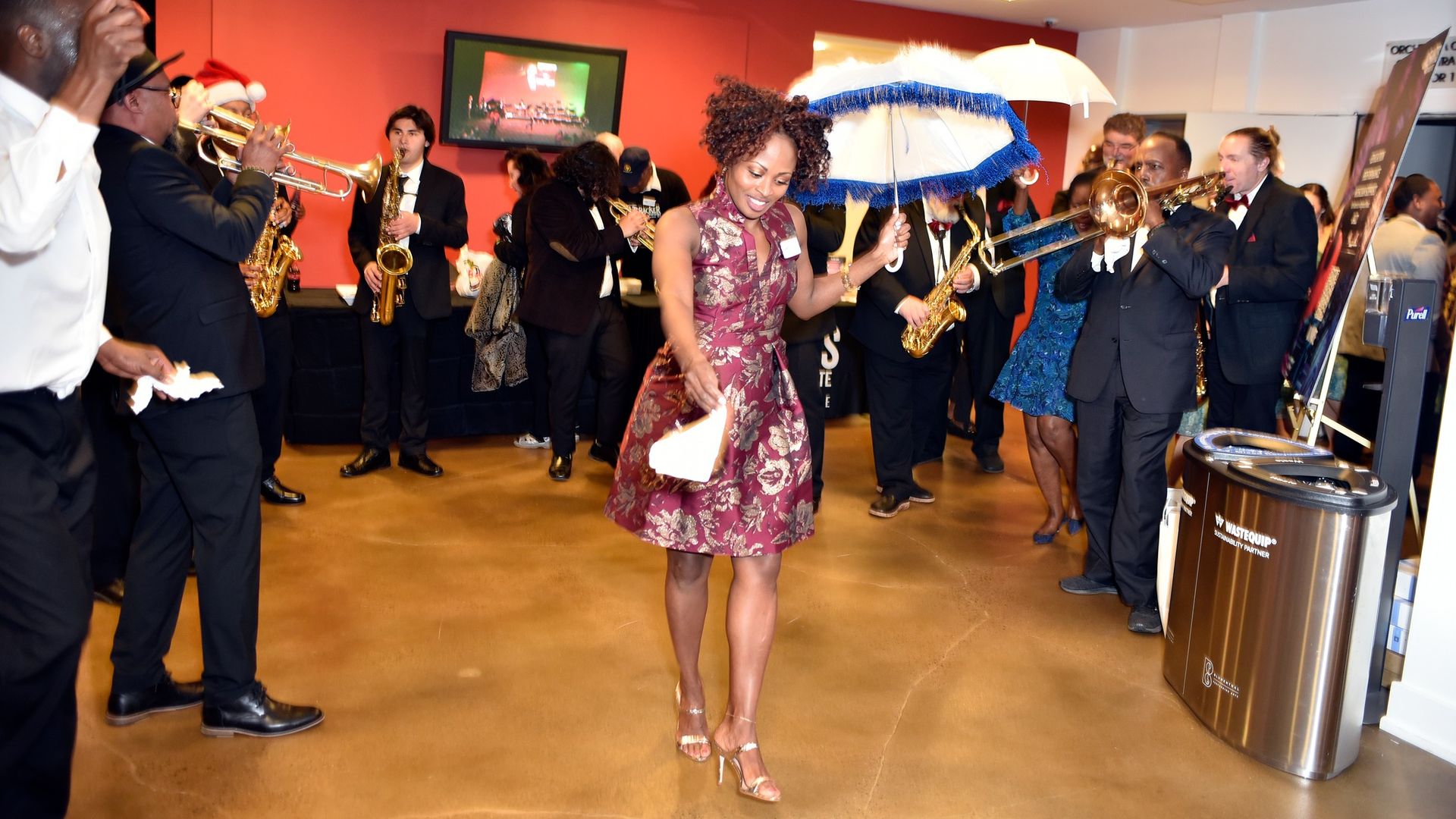 A lively indoor scene with a woman in a maroon floral dress dancing with a white umbrella, surrounded by musicians playing brass instruments in formal attire against a red and white wall.