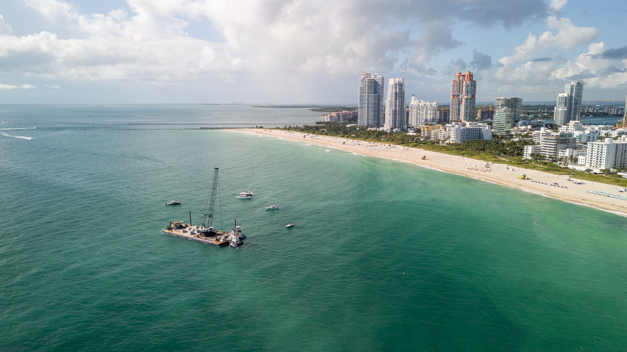 Aerial view of a turquoise ocean with a floating construction platform and boats near a sandy beach lined with tall buildings under a partly cloudy sky.