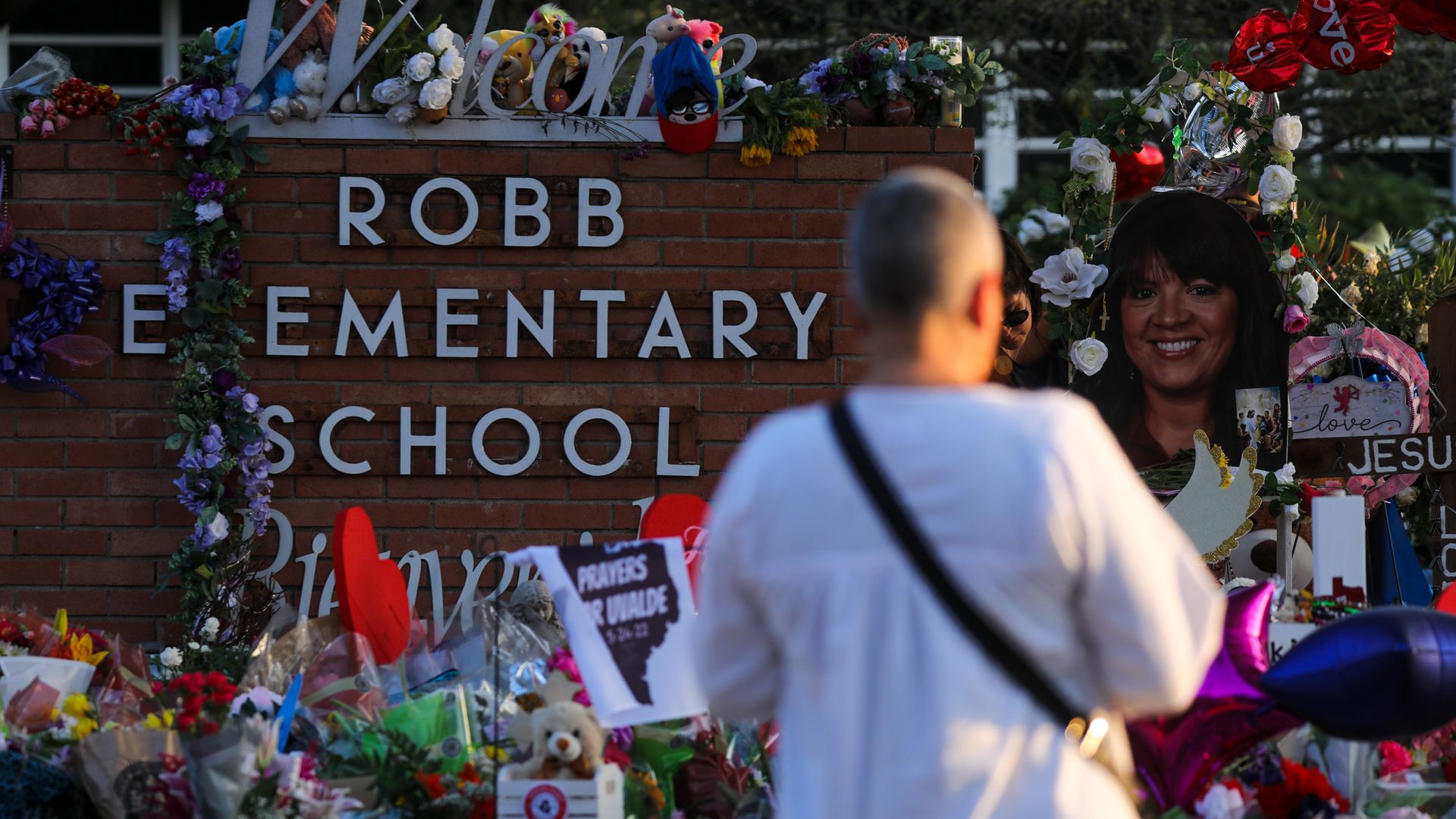 Photo of a person standing in front of a brick sign that says "Robb Elementary School" and is shrouded by flowers and signs