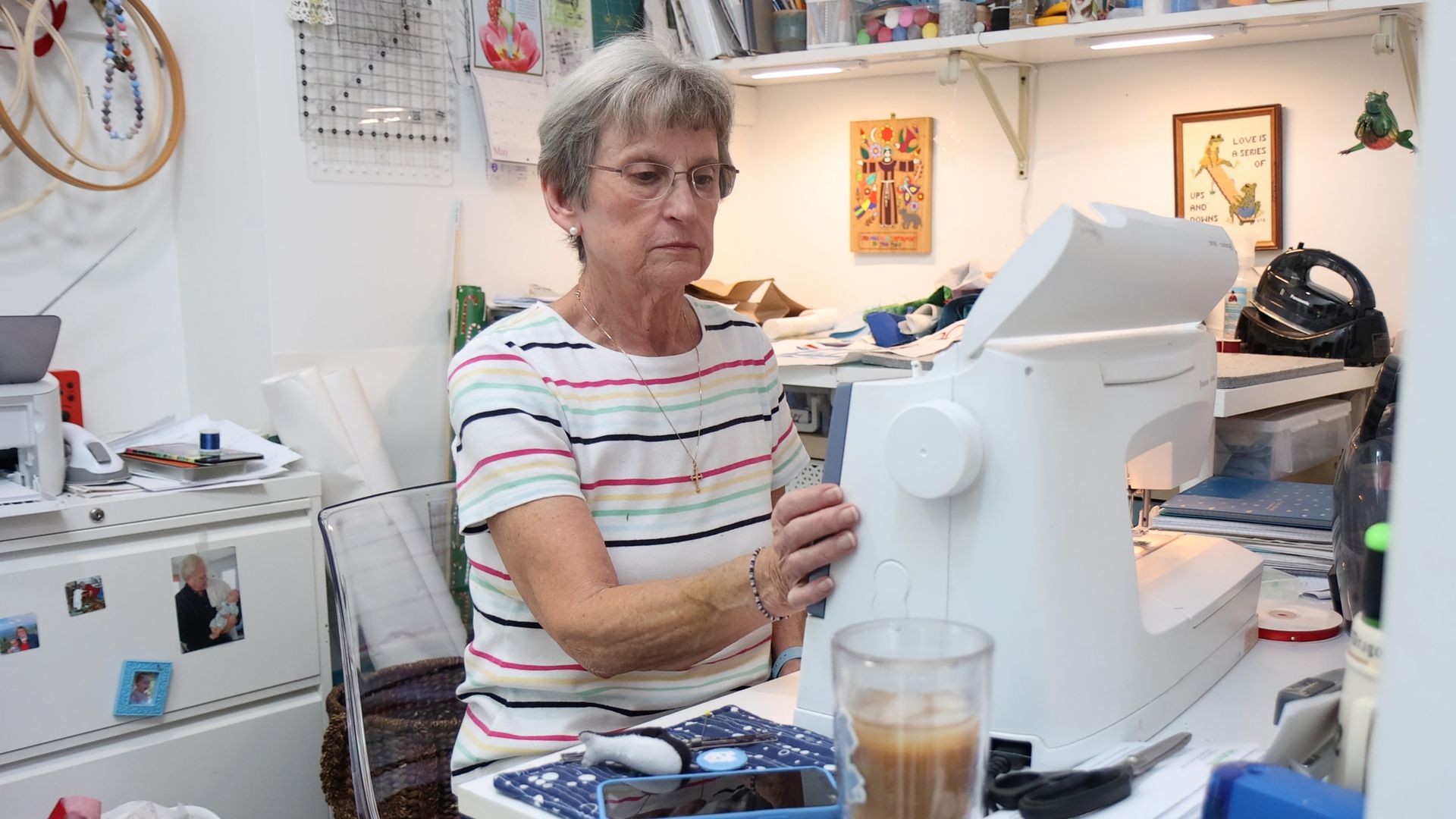 An older woman with short gray hair and glasses sits at a sewing machine in a bright, cluttered craft room, wearing a striped shirt, surrounded by fabric, papers, posters, and drafting tools.