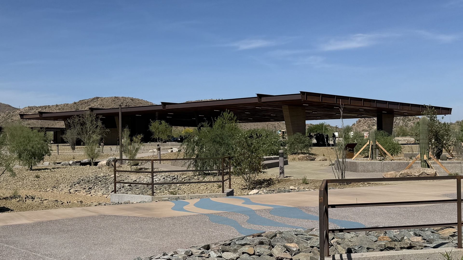 Desert park featuring a long, brown metal pavilion under a blue sky. Sparse trees, cacti, and rocks surround a blue-waved path; brown railings border the foreground.