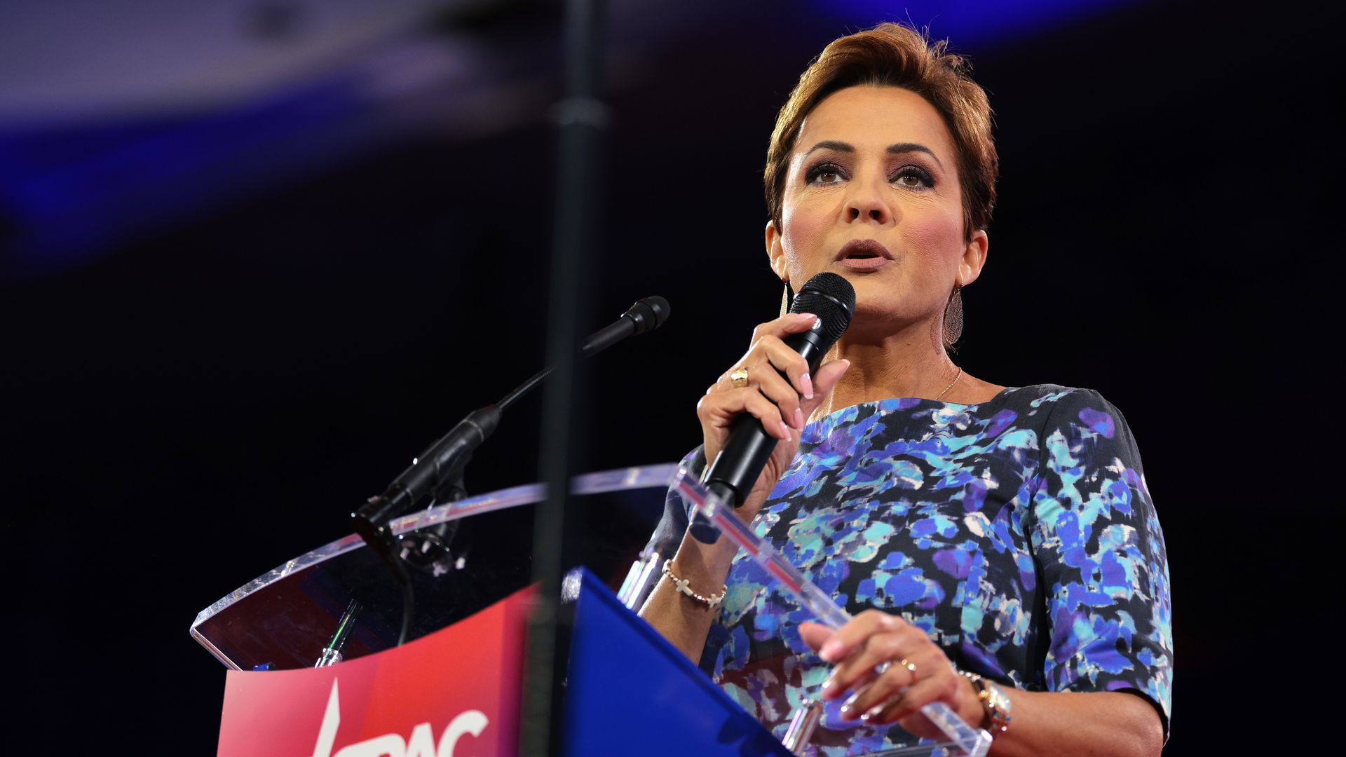 A woman in a dress with a blue and purple floral pattern against a black background holds a microphone to her mouth while standing at a lectern with a logo showing the letters "CPAC" next to a star. 