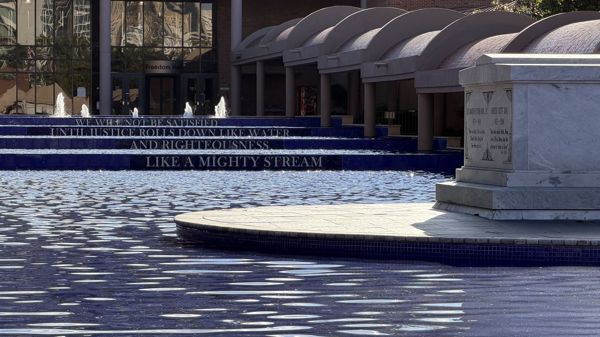 Tiered fountain steps display the quote, "We will not be satisfied until justice rolls down like waters and righteousness like a mighty stream." A marble monument marking the graves of Dr. Martin Luther King, Jr., and Coretta Scott King sits to the right; a building behind.