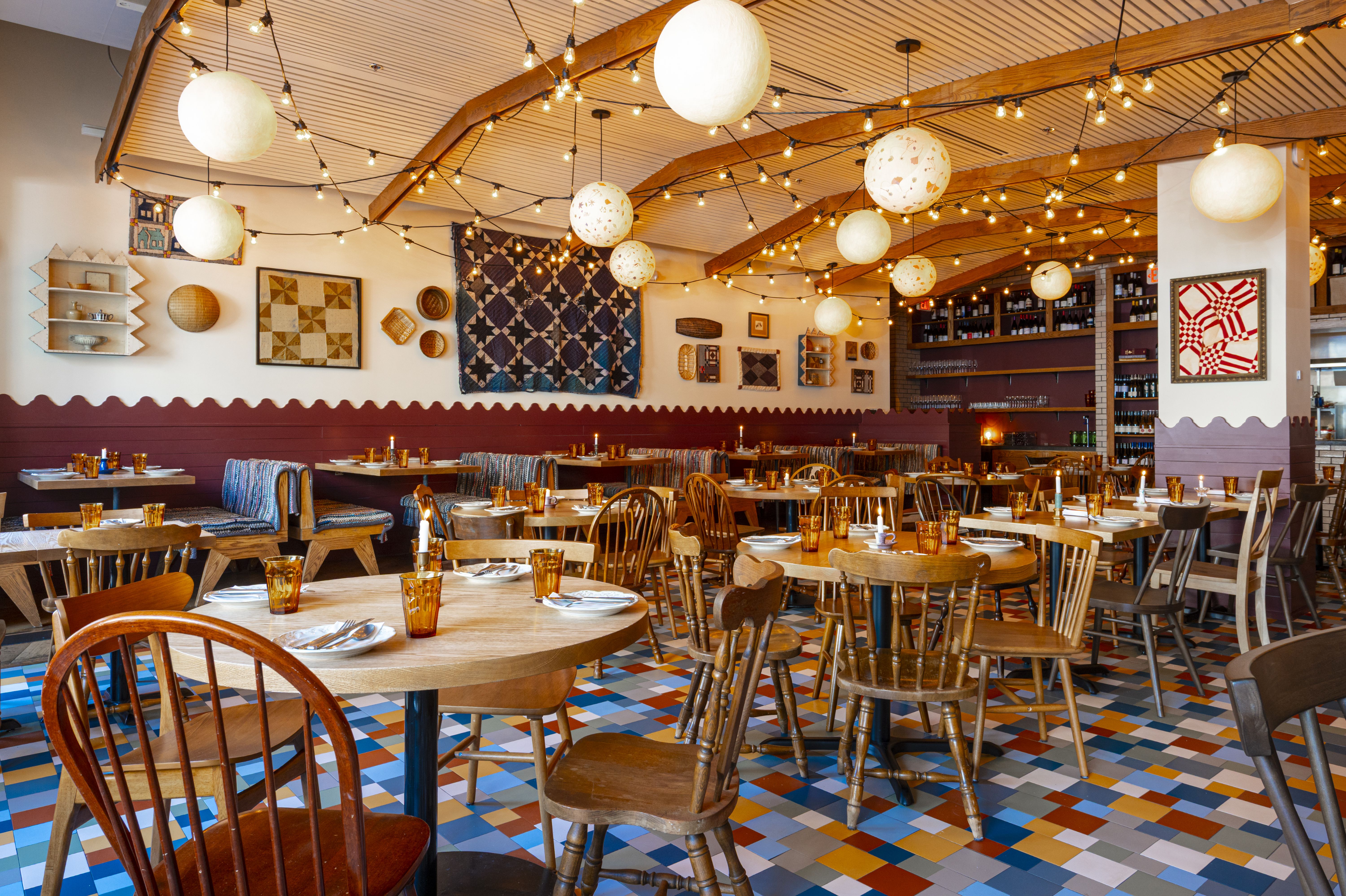 Cozy, eclectic restaurant interior with wooden tables and mixed chairs, amber glassware, warm string lights, large round lights, patchwork quilts on maroon walls, and a colorful tiled floor.