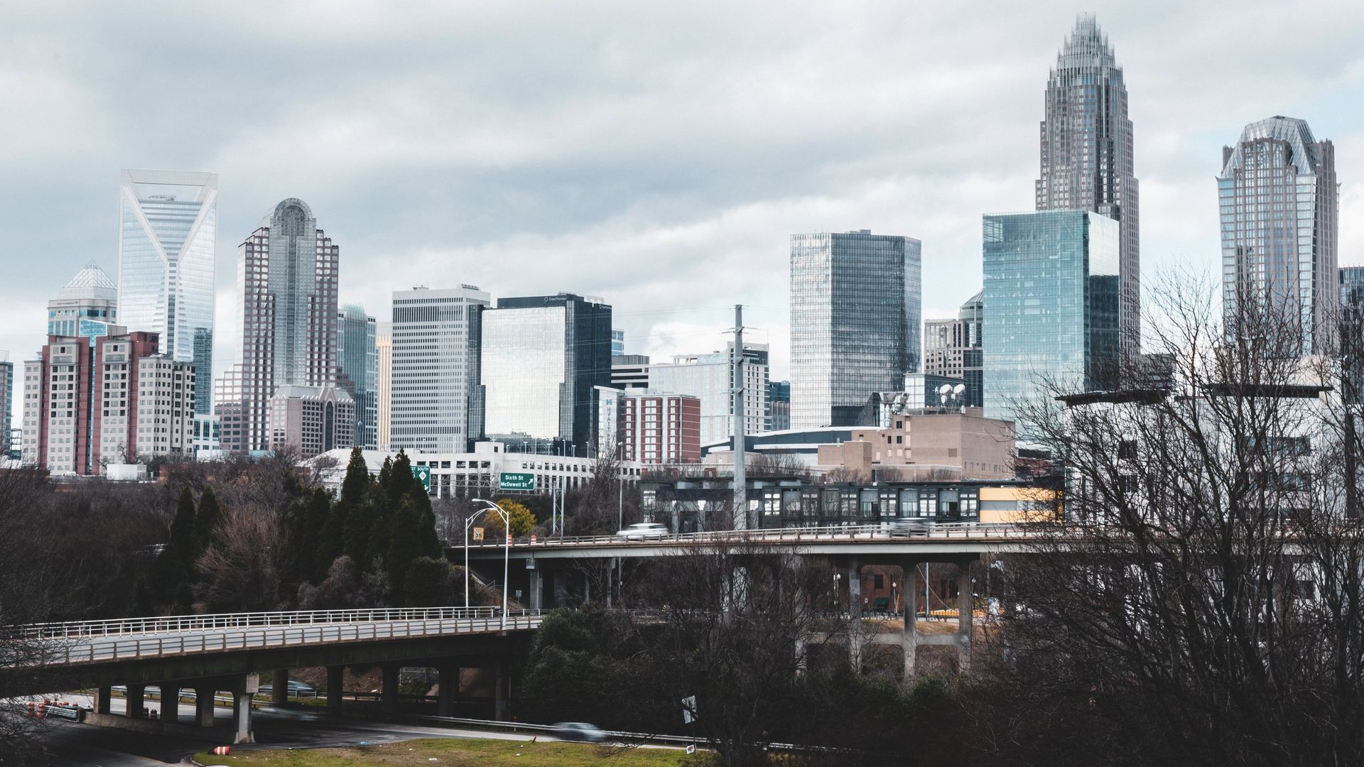 City skyline with glass skyscrapers, a curved elevated highway in the foreground, leafless trees, and a gray, overcast sky.