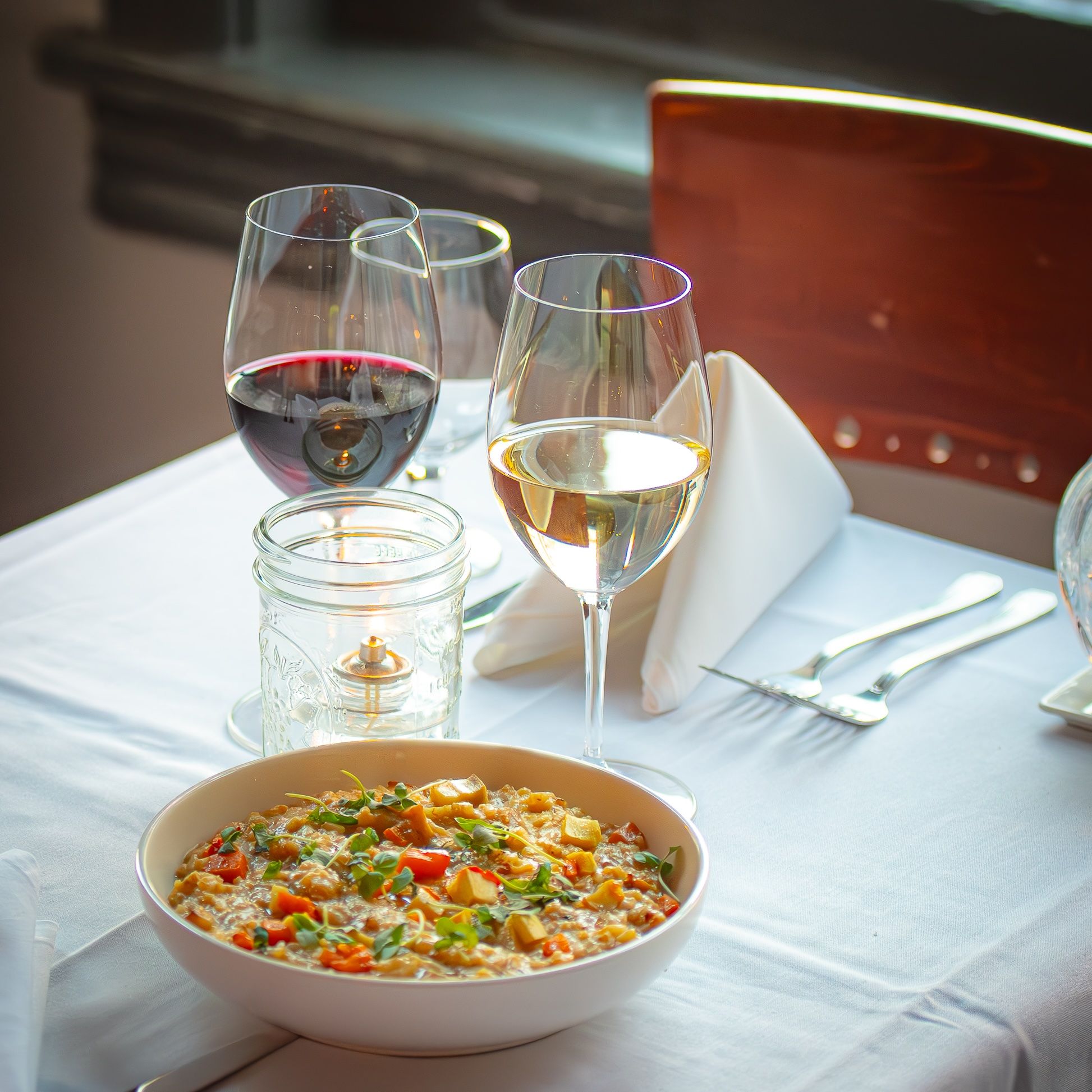 A white tablecloth set with a bowl of colorful vegetable risotto, two wine glasses filled with red and white wine, a candle in a glass jar, and folded white napkins in a restaurant setting.