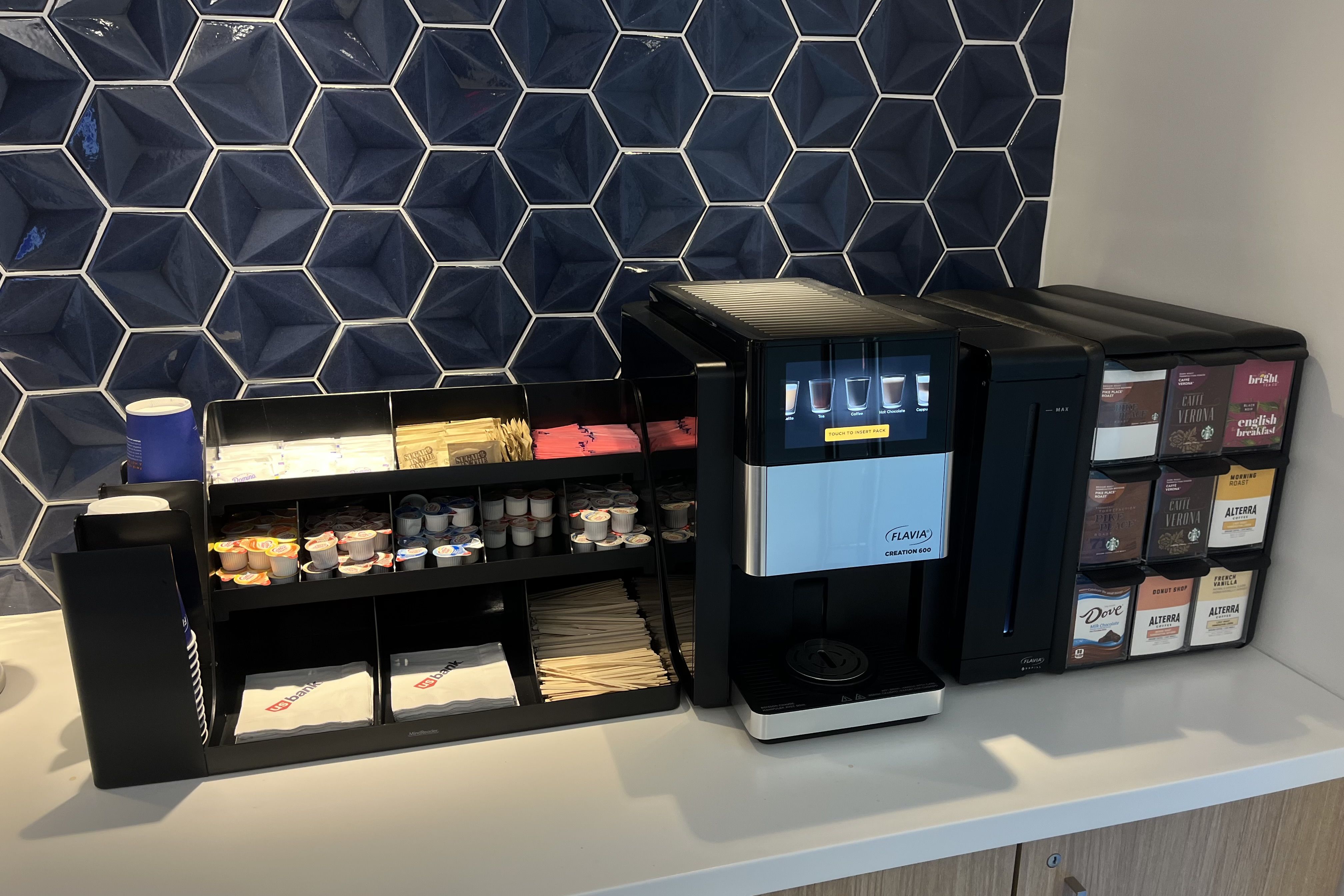 Coffee station with a Flavia Creation 600 machine, various creamers and sugar packets, wooden stirrers, napkins, and blue hexagonal tile background.