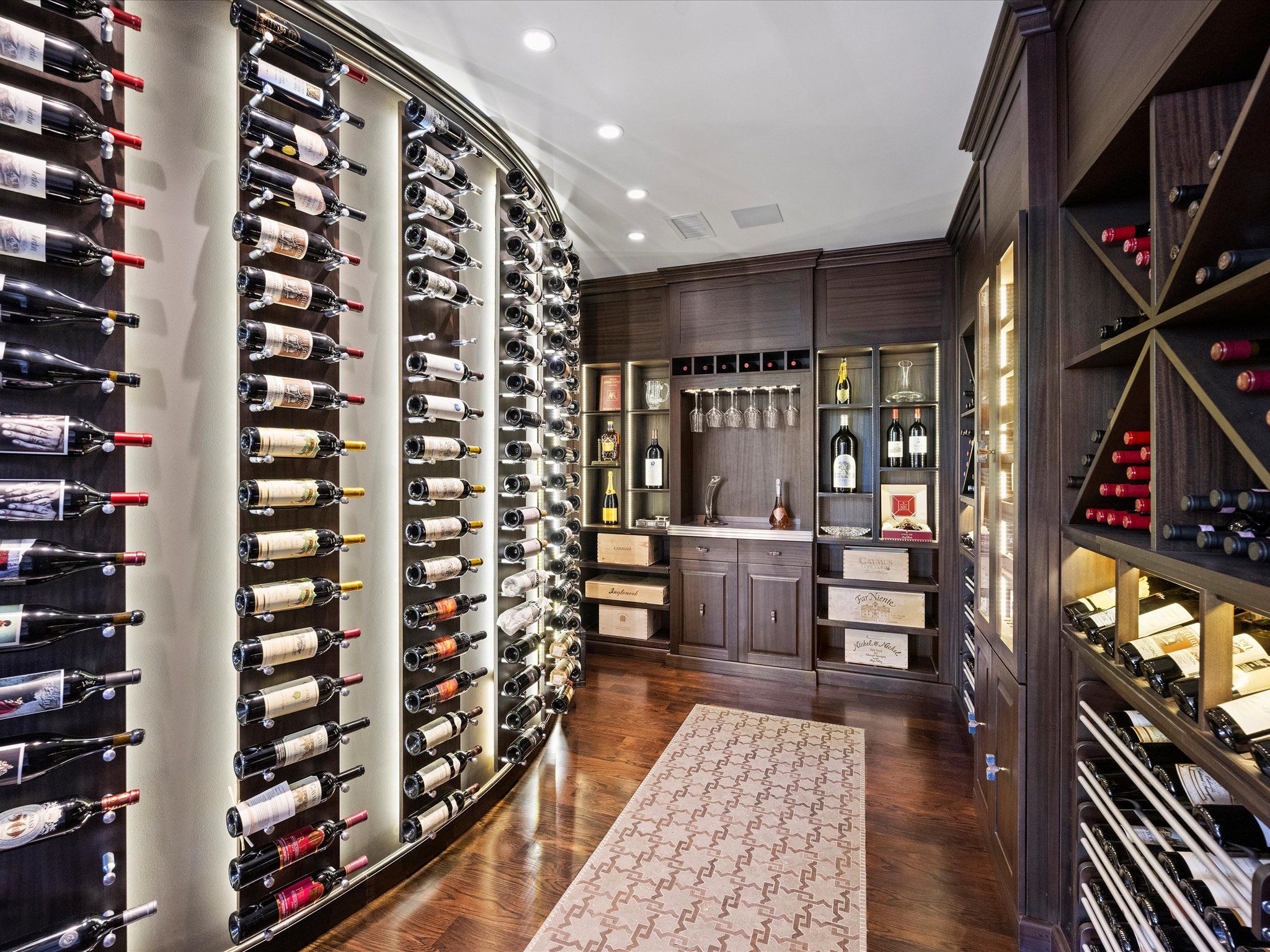 Modern wine cellar with wood flooring and cabinetry, curved backlit metal wall rack holding many bottles, shelves with wine boxes and glassware, and a patterned beige rug on the floor.