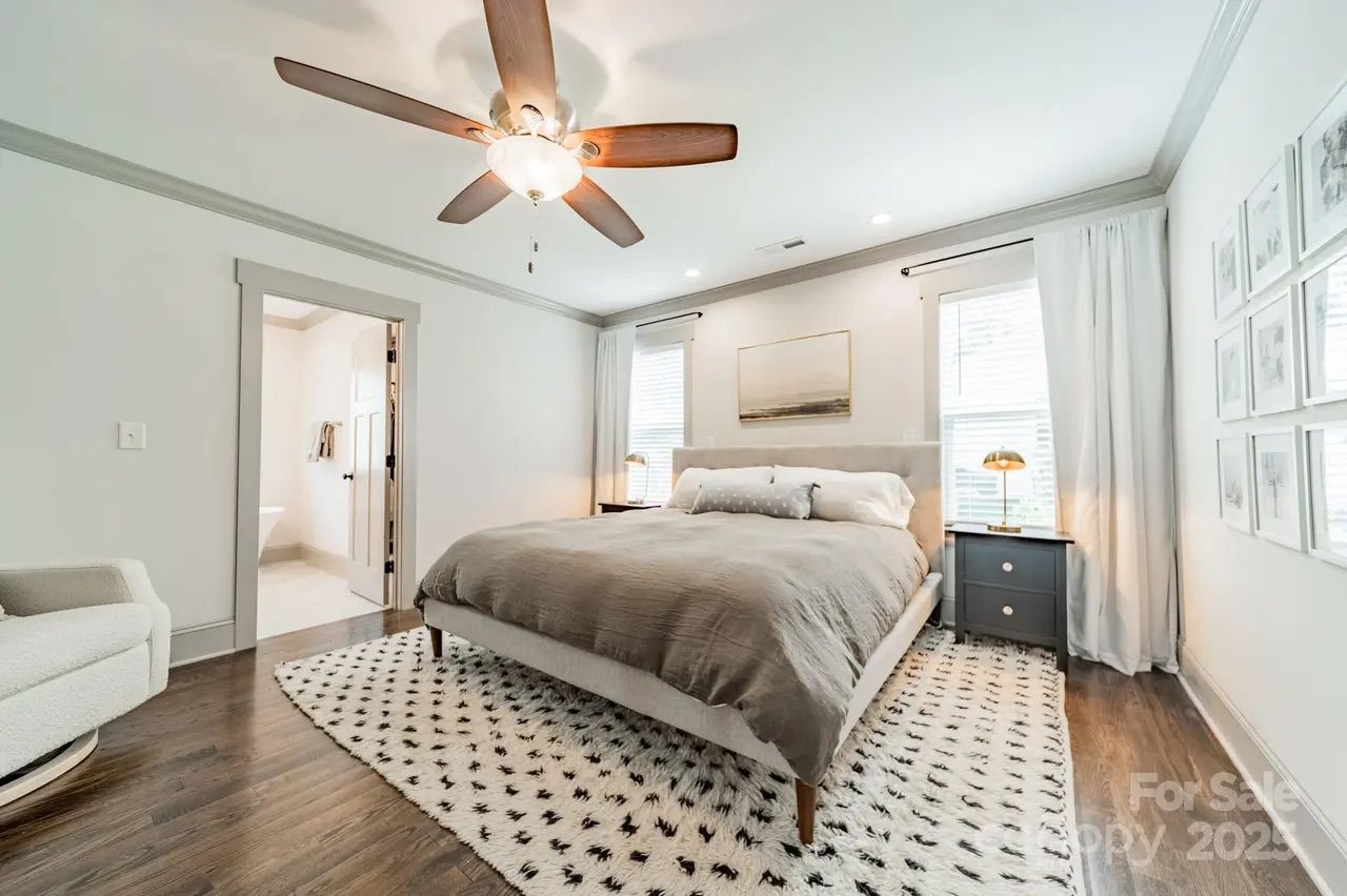 Bright bedroom with gray bedspread, beige upholstered bed frame, patterned rug, wooden floors, ceiling fan, and two dark nightstands with gold lamps by windows with white curtains.