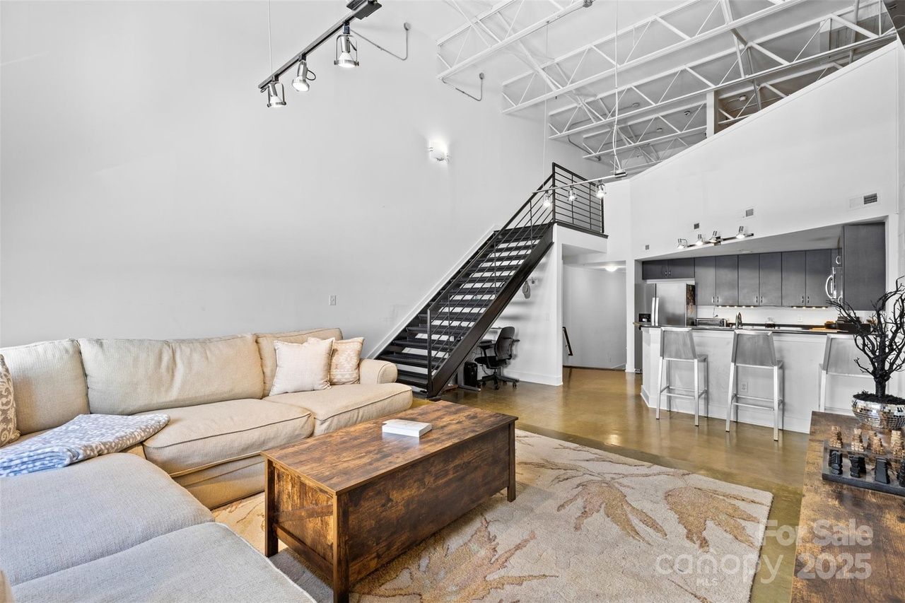 Open living space with beige sectional sofa, wooden coffee table, modern kitchen with black cabinets and bar stools, and black metal staircase leading to loft.