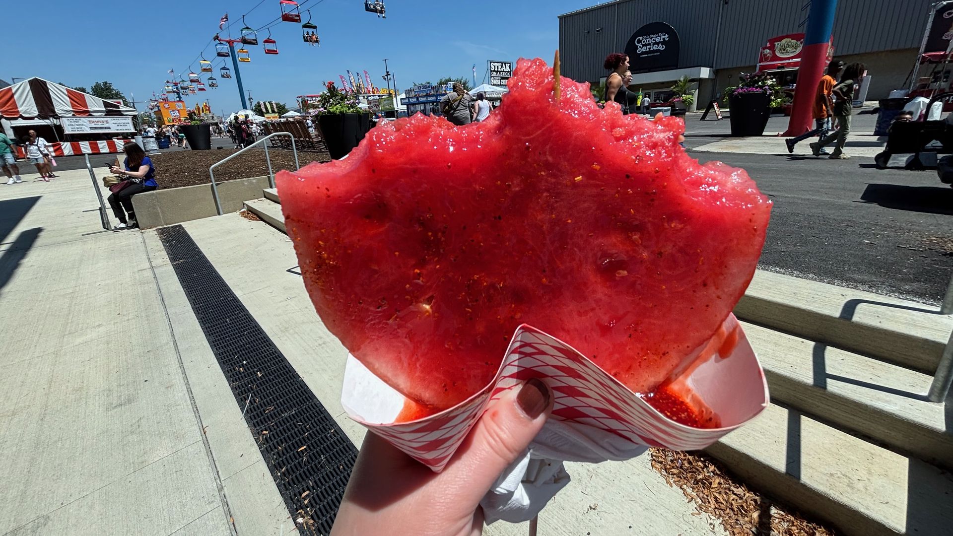 Hand holding a slice of half-eaten watermelot on a stick at the state fair with blue sky, people, tents, and colorful sky lift chairs in the background.