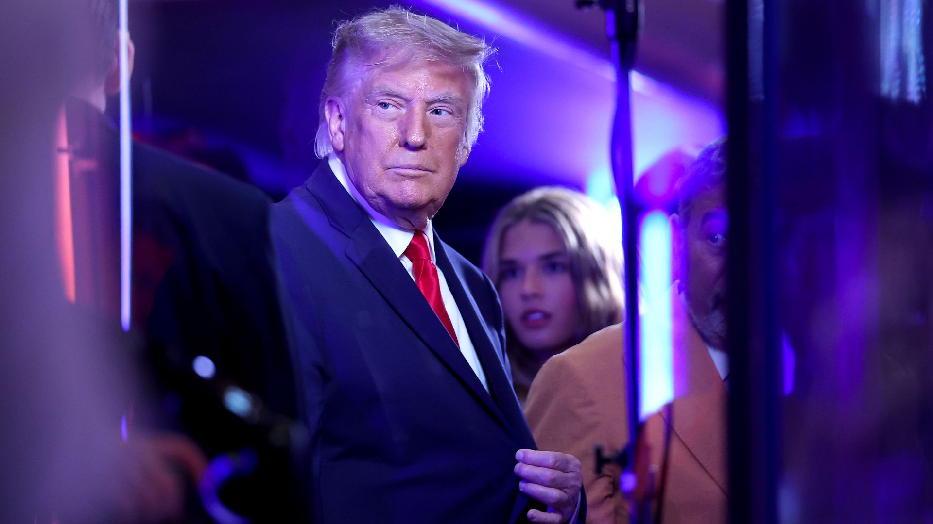 President Trump, a man in a dark suit and red tie stands near a glass barrier in a blue and purple-lit venue, with a blurred crowd in the background and bright lights reflecting on his face.