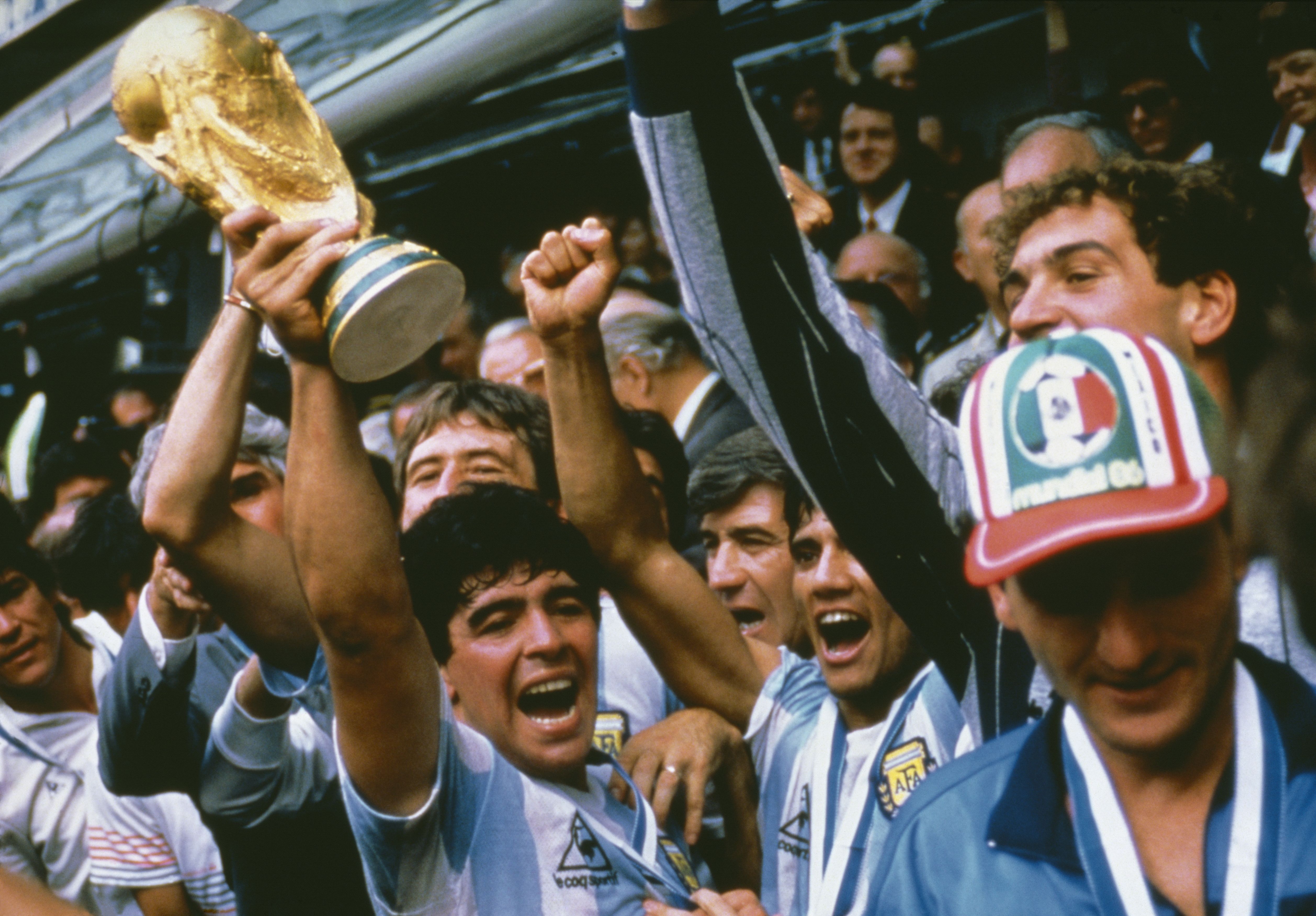 Argentinian soccer players in blue and white jerseys celebrating and lifting the golden FIFA World Cup trophy, surrounded by cheering crowd and officials.