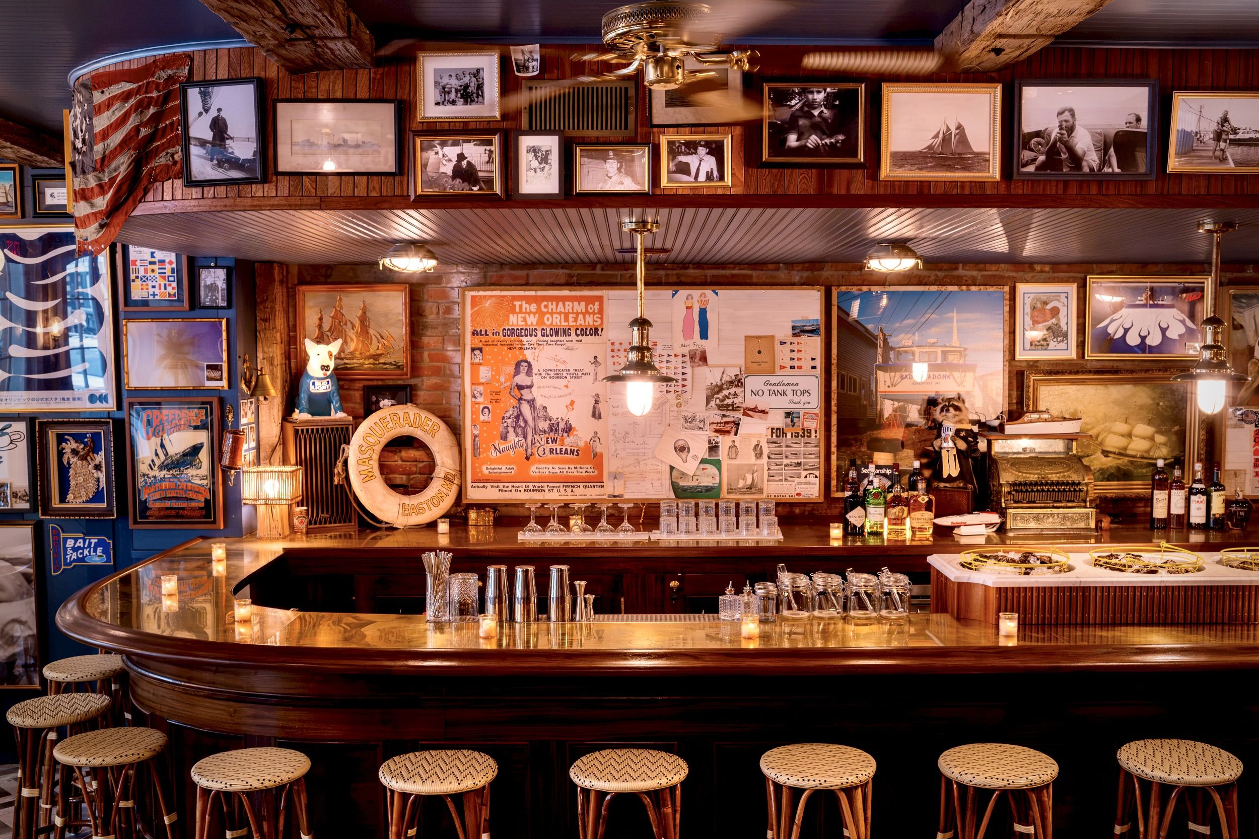 Warm, vintage bar with a curved wooden counter and wicker stools. Brick walls are lined with black-and-white photos and posters; an American flag drapes along the ceiling, illuminated by warm lights.