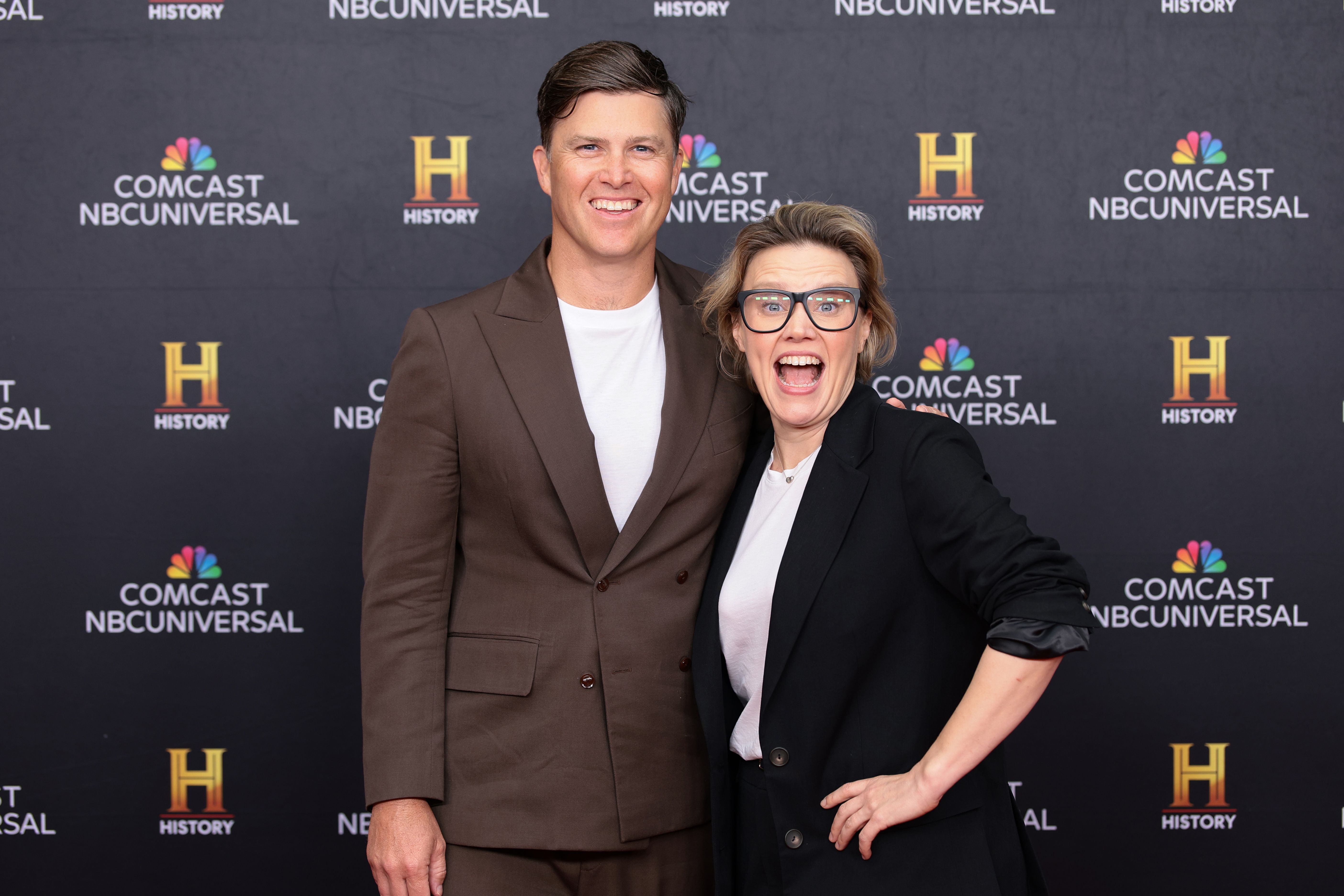Two smiling people pose on a black NBCUniversal/History backdrop; a man in a brown suit stands beside a laughing woman in a black blazer and white top.