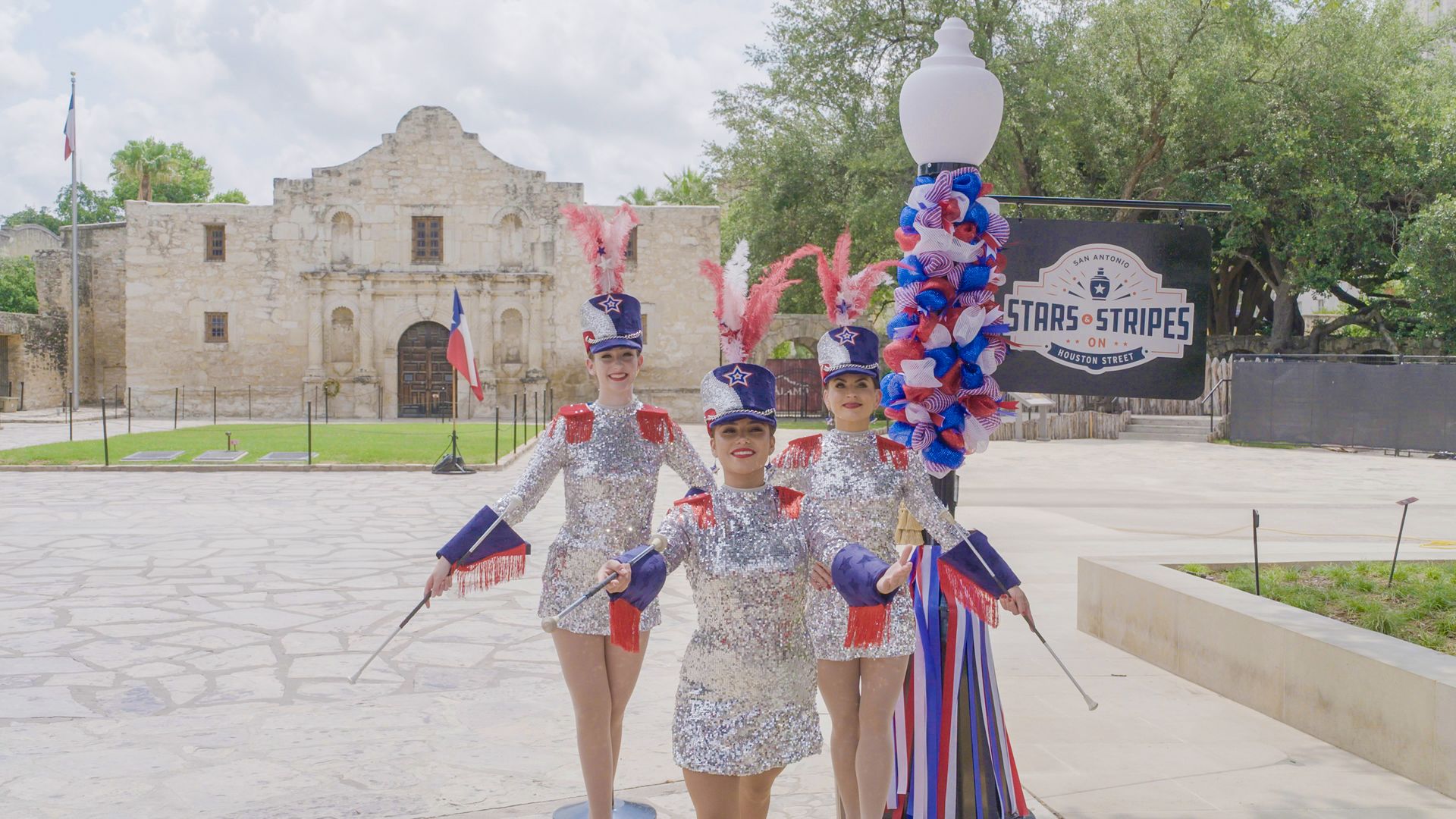 Dancers dressed in red, white and blue outfits and hats pose in front of the Alamo.