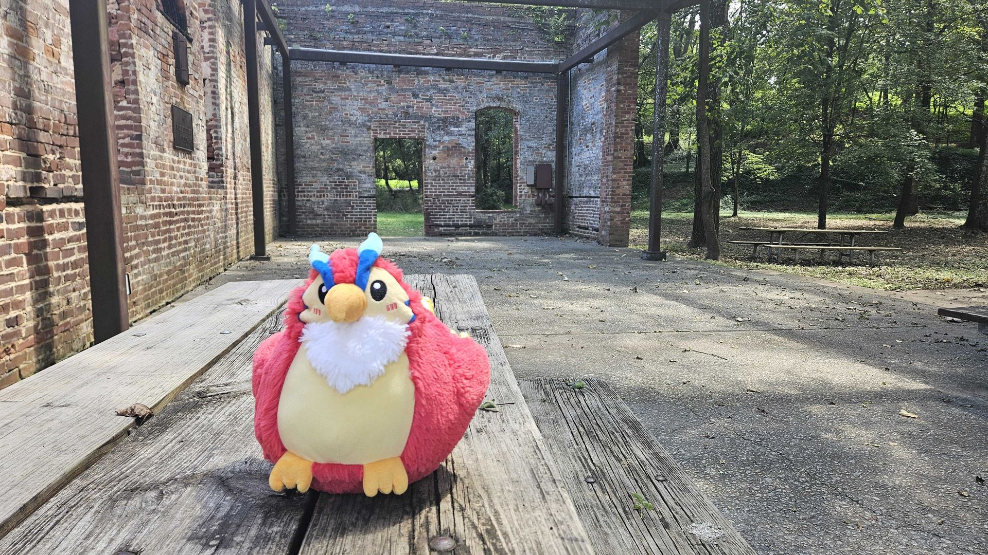 Plush red bird with blue horns and yellow beak sitting on wooden picnic table outdoors near brick ruins and green trees on a sunny day.