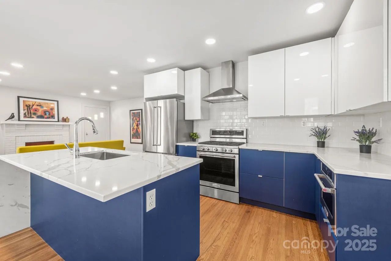 Modern kitchen with blue lower cabinets and white upper cabinets, marble countertops, stainless steel appliances, wood floor, and a kitchen island with a sink under white recessed lighting.