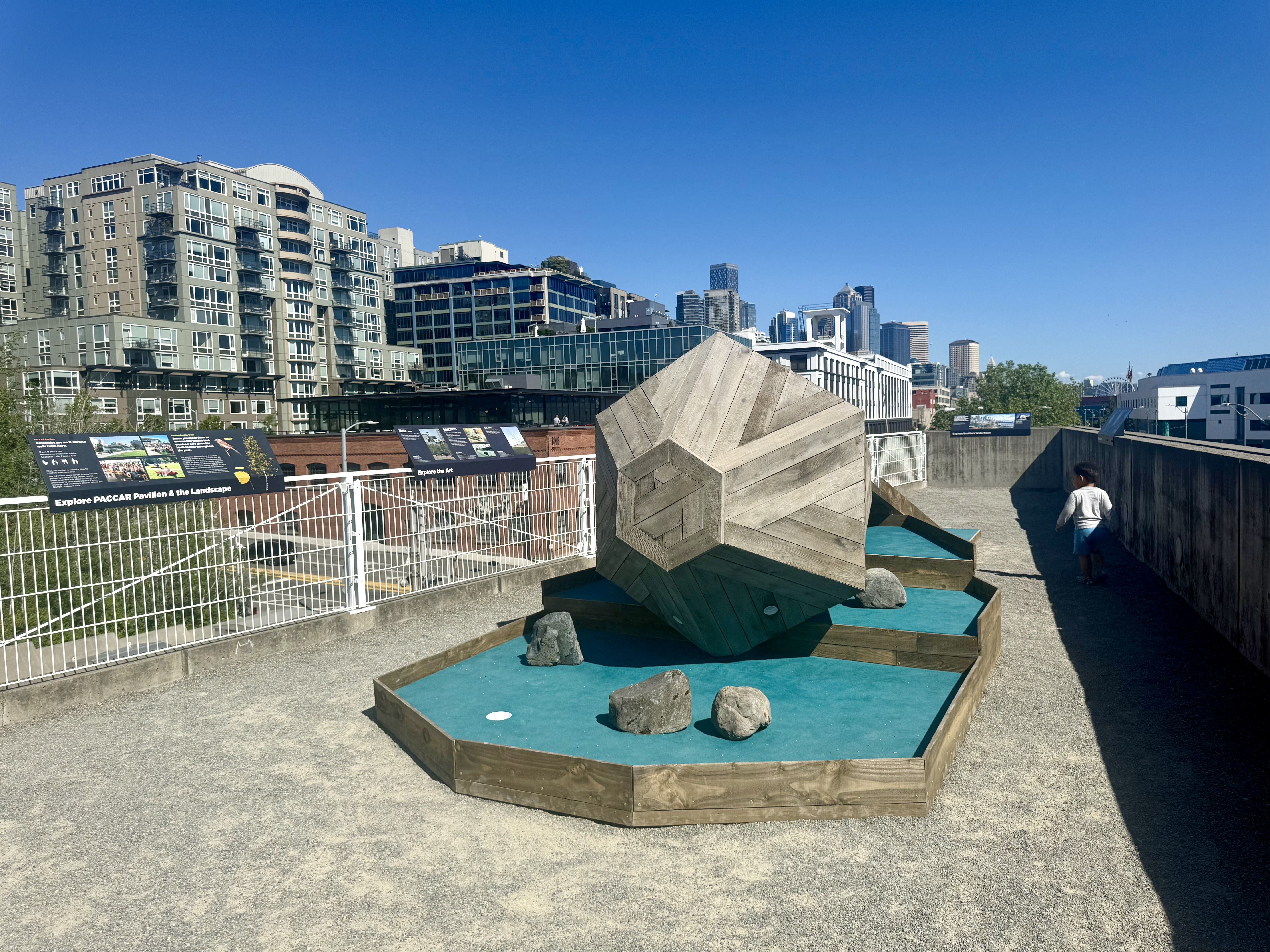 City skyline of modern buildings by a waterfront beneath a clear blue sky. In the foreground, a large geometric wooden sculpture sits on turquoise putt putt area as child walks nearby.