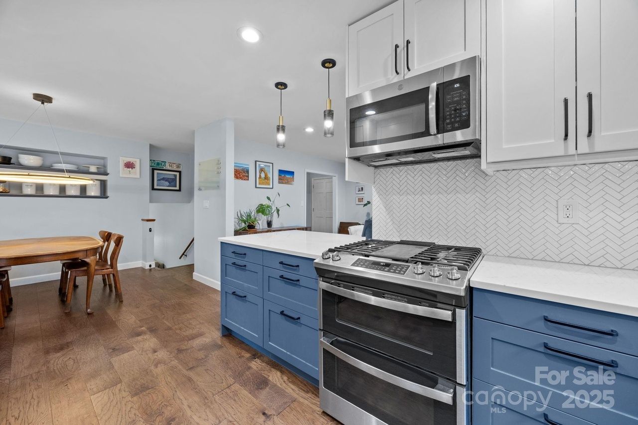 Modern kitchen with blue lower cabinets, white upper cabinets, stainless steel stove and microwave, white herringbone tile backsplash, wooden dining table with chairs, and hardwood floor.
