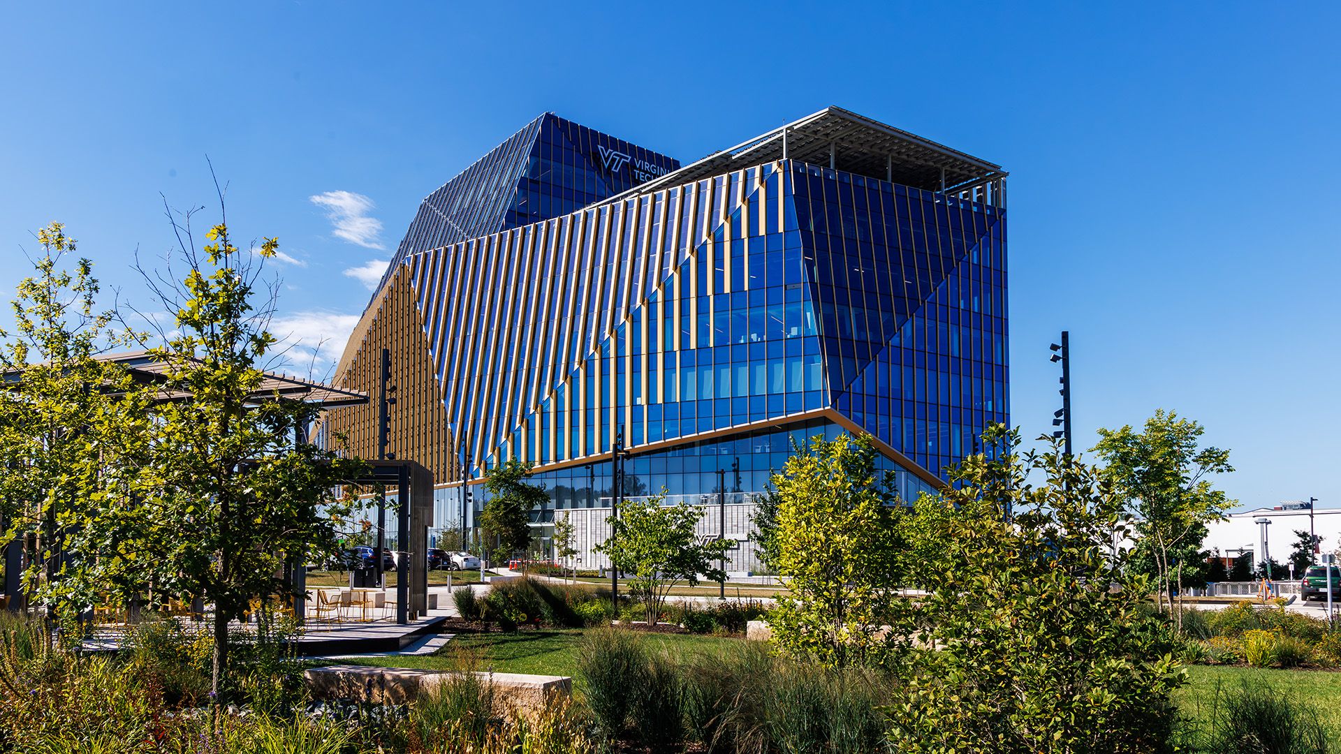 Modern blue glass building with diagonal panels, set against a clear blue sky. Green landscaping frames the foreground; Virginia Tech logo visible on the upper facade.