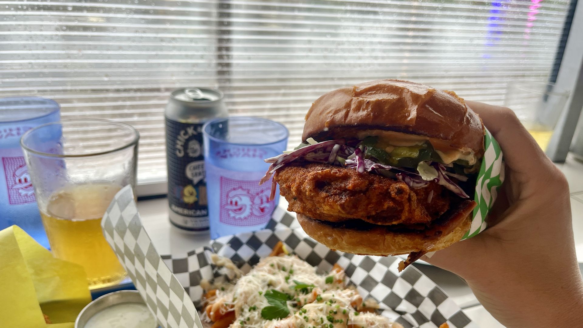 Hand holding a fried chicken sandwich with pickles and purple cabbage on a toasted bun, with fries, a beer and two blue cups with chicken logos on a table by a window.