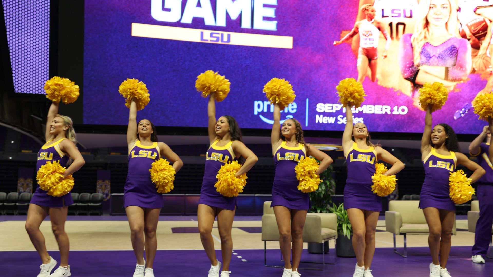 Six cheerleaders seen on a court in front of a large screen displaying a promo for "The Money Game." 