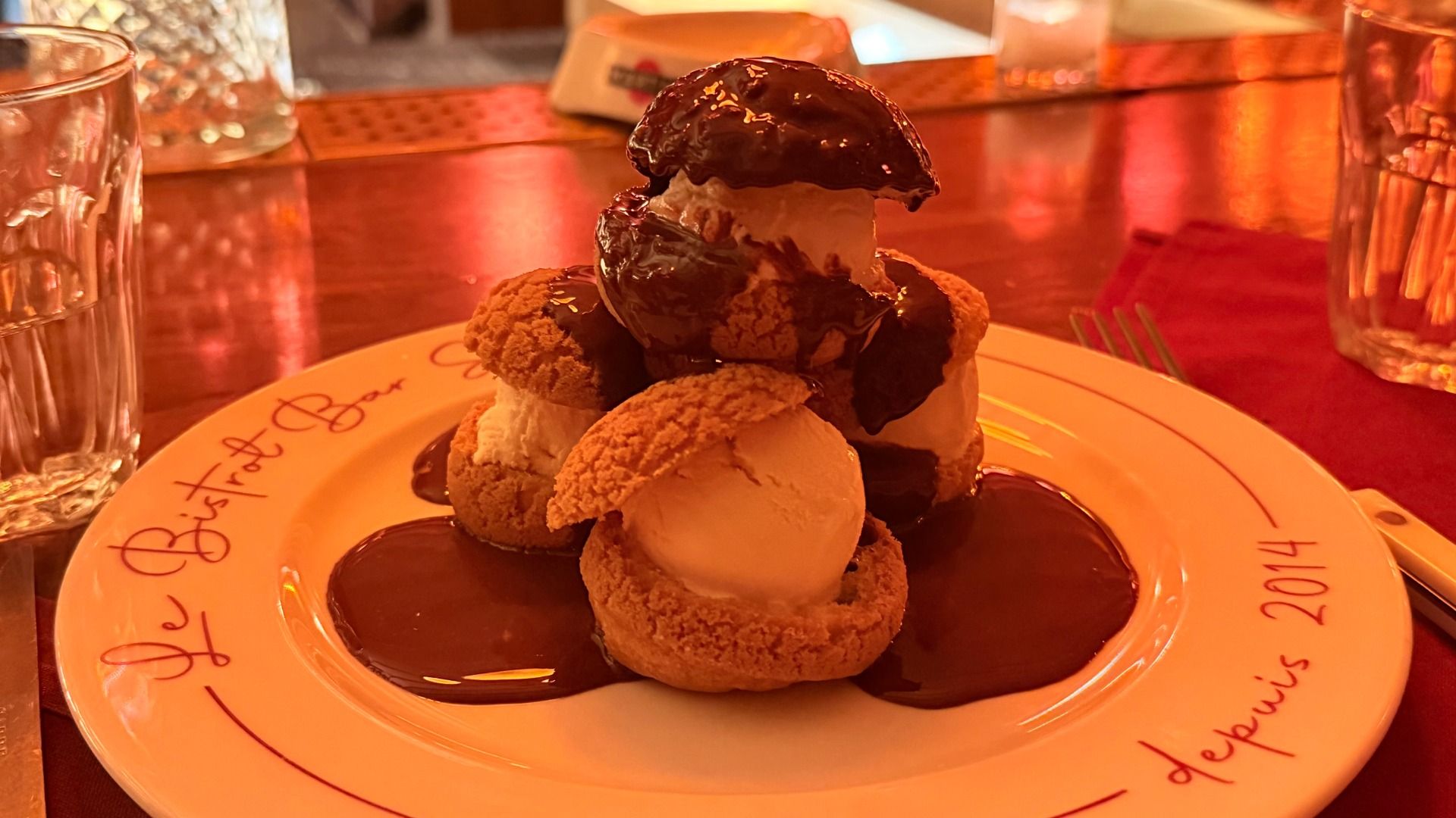 Plate of ice cream-filled pastries topped with dark chocolate sauce on a white plate with red script, surrounded by glasses and red fabric on a wooden table.