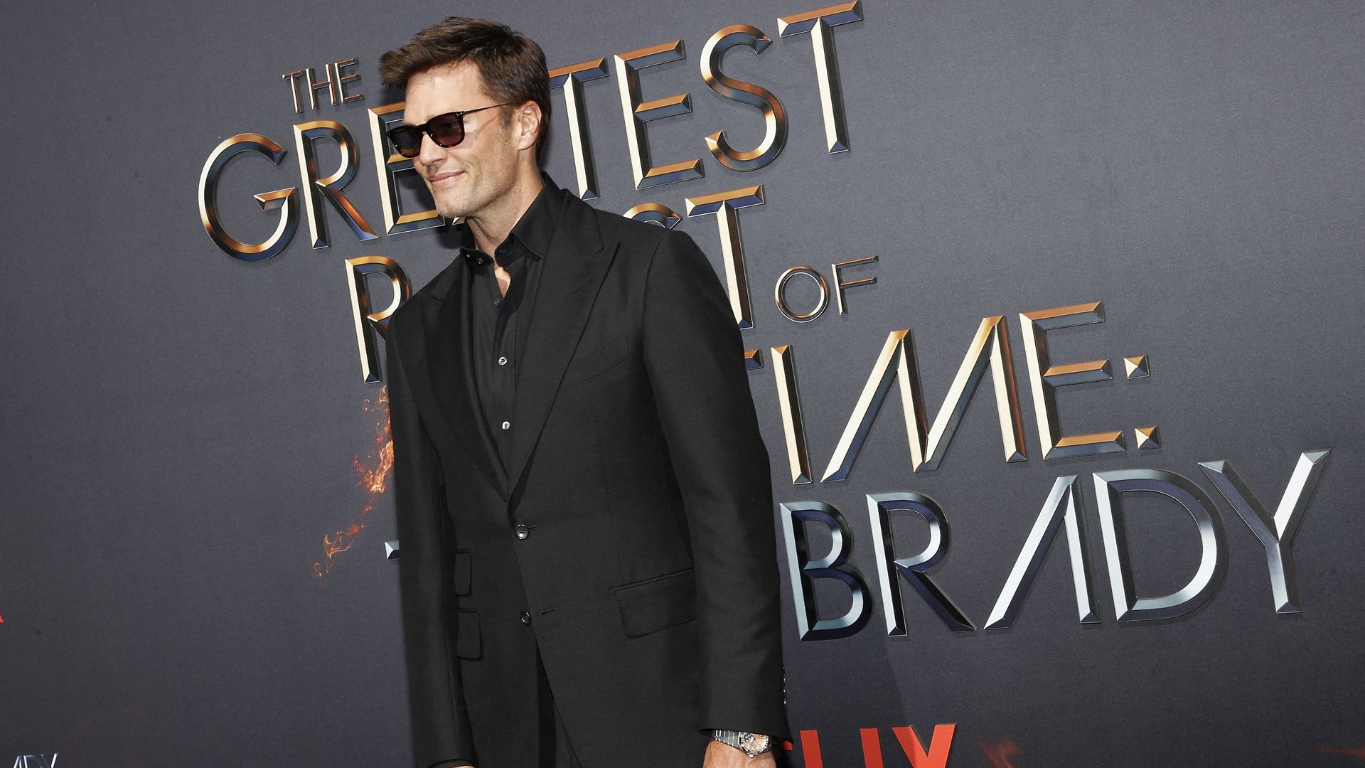 A person in a black suit and sunglasses stands in front of the logo for a netflix show