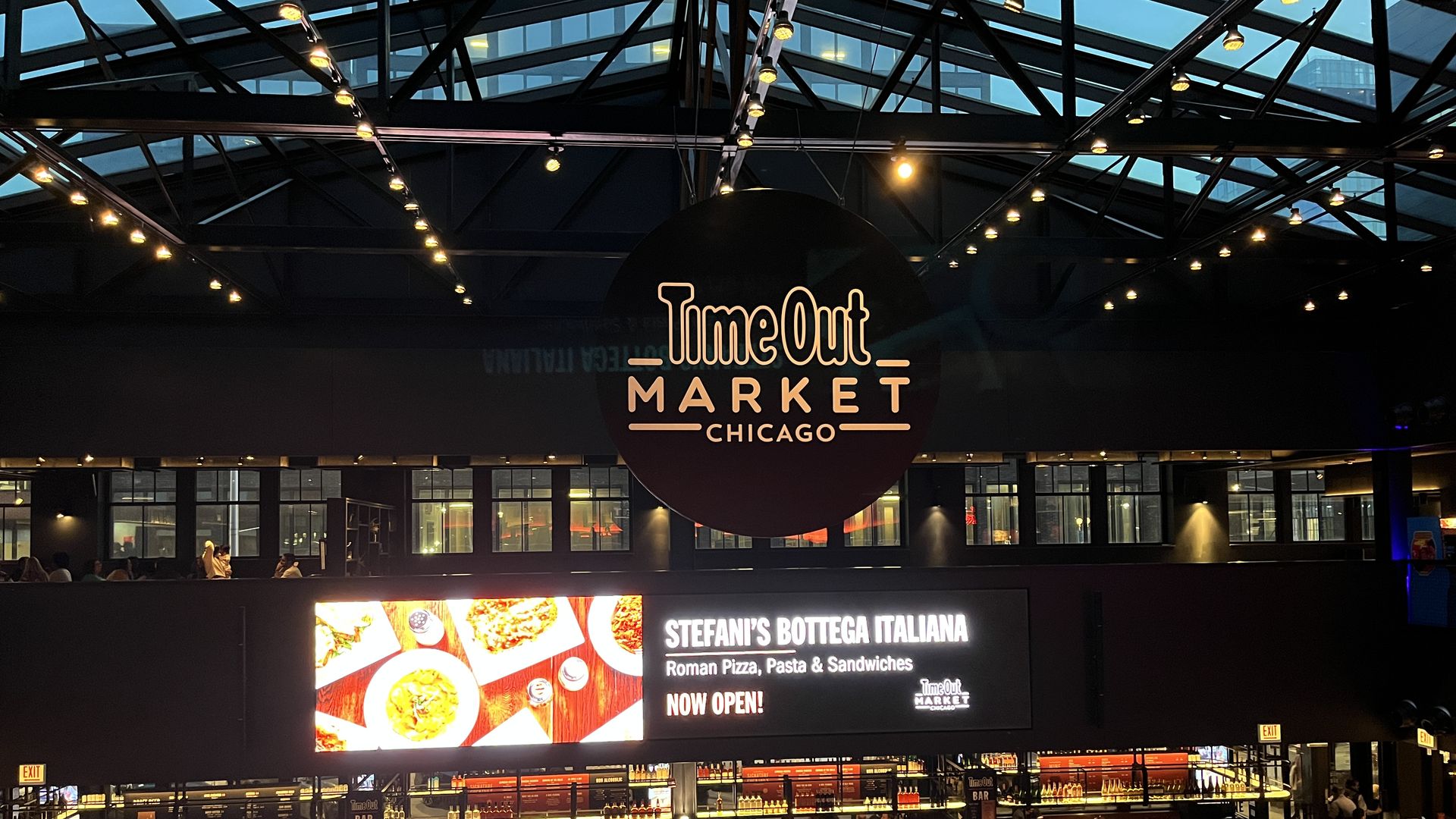 Indoor view of Time Out Market Chicago with hanging black circular sign, lit ceiling lights, digital sign for Stefani's Bottega Italiana, and people dining at wooden tables below.