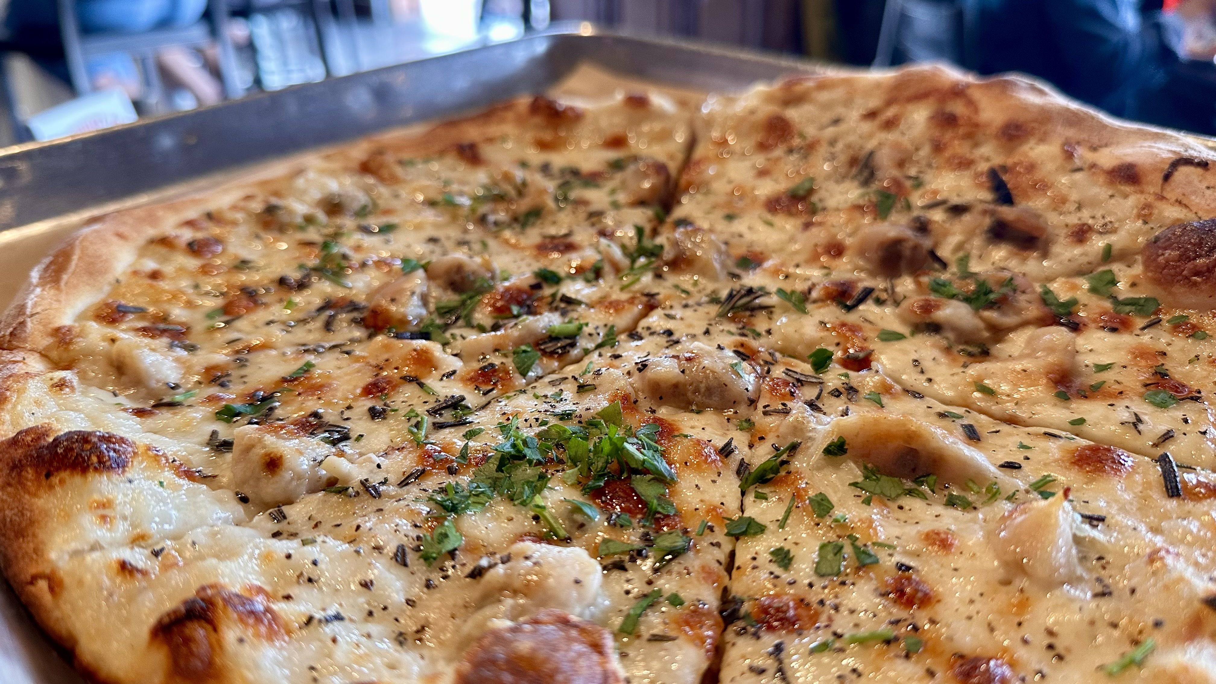 Close-up of a freshly baked pizza with melted cheese, herbs, black pepper, and chunks of clam on a metal tray in a restaurant setting.