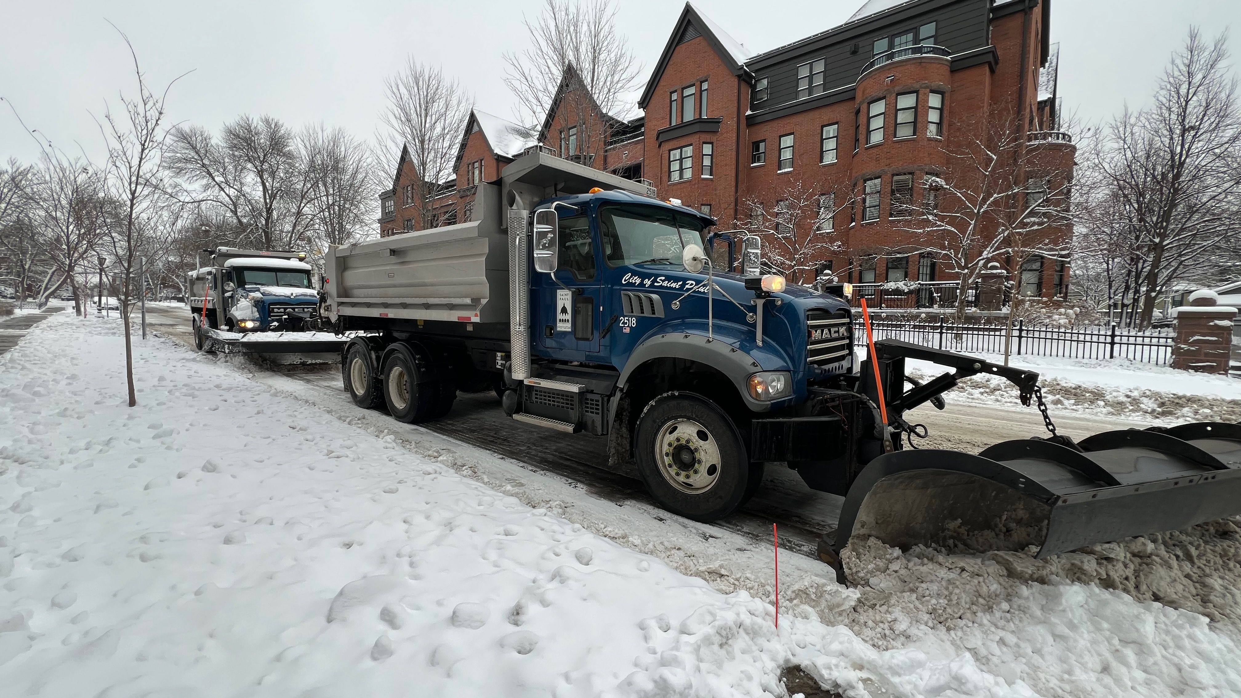A snowplow pushing snow to the curb in front of a brown brick apartment building