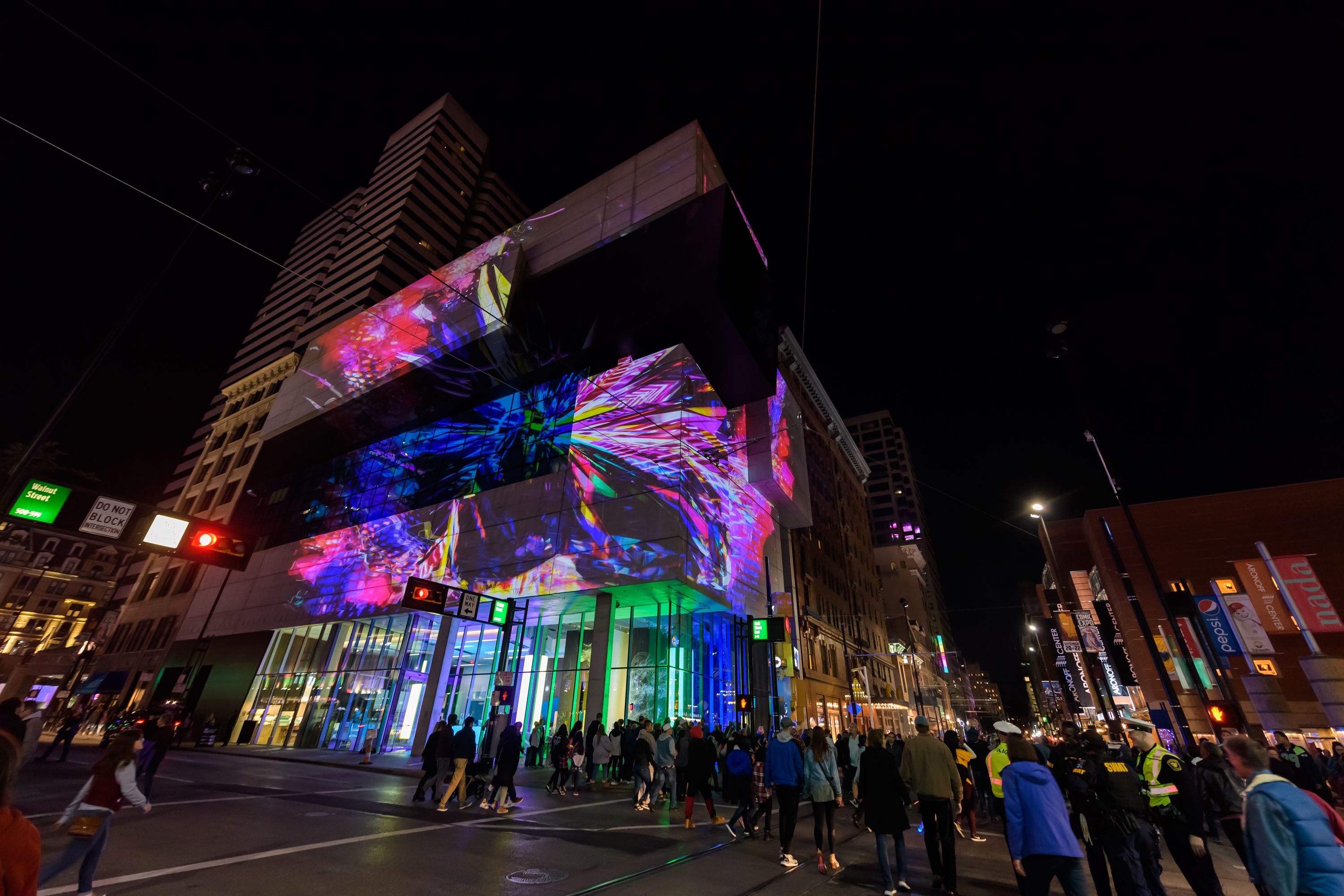 Colorful lights and patterns are projected onto the exterior of a modern building at night, with crowds of people walking by.