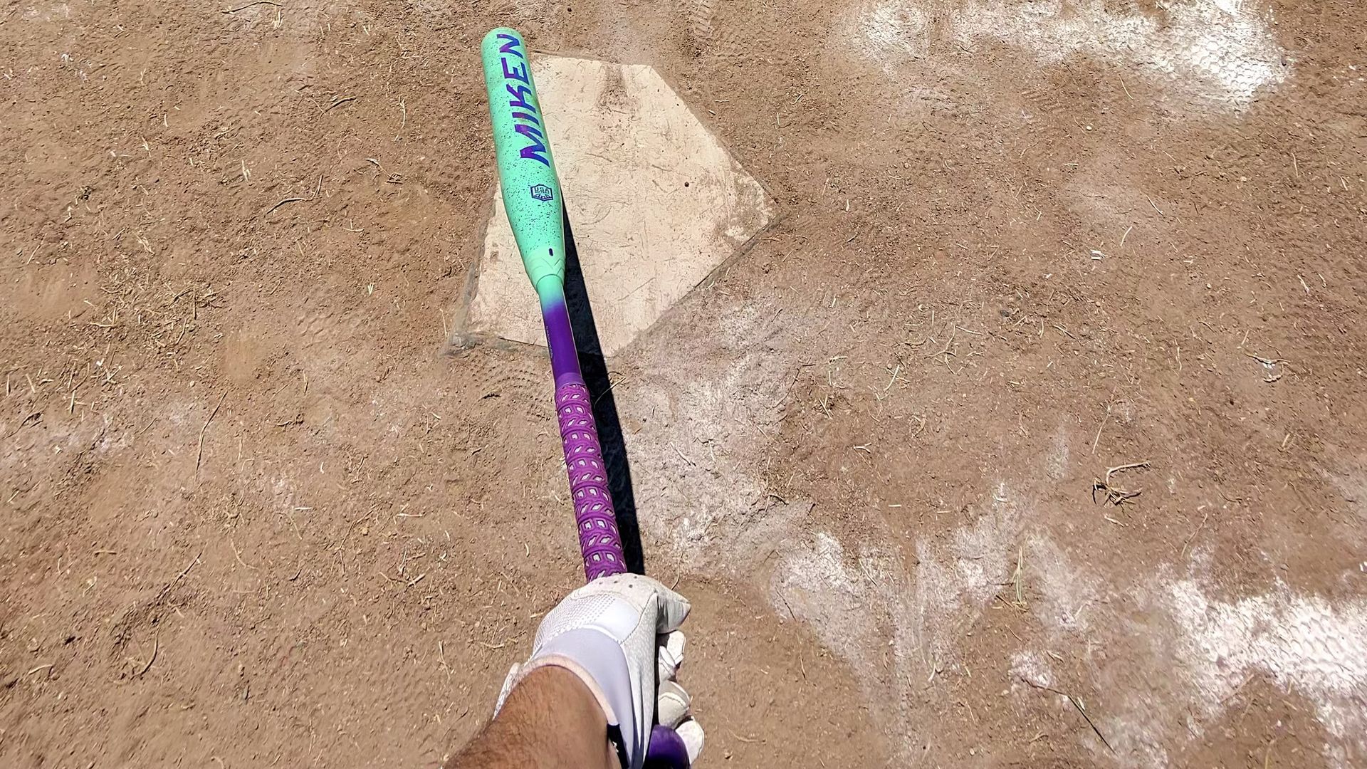 View from a batter's perspective holding a turquoise and purple Miken bat over a home plate on a dry, dusty baseball field with faded white chalk lines.